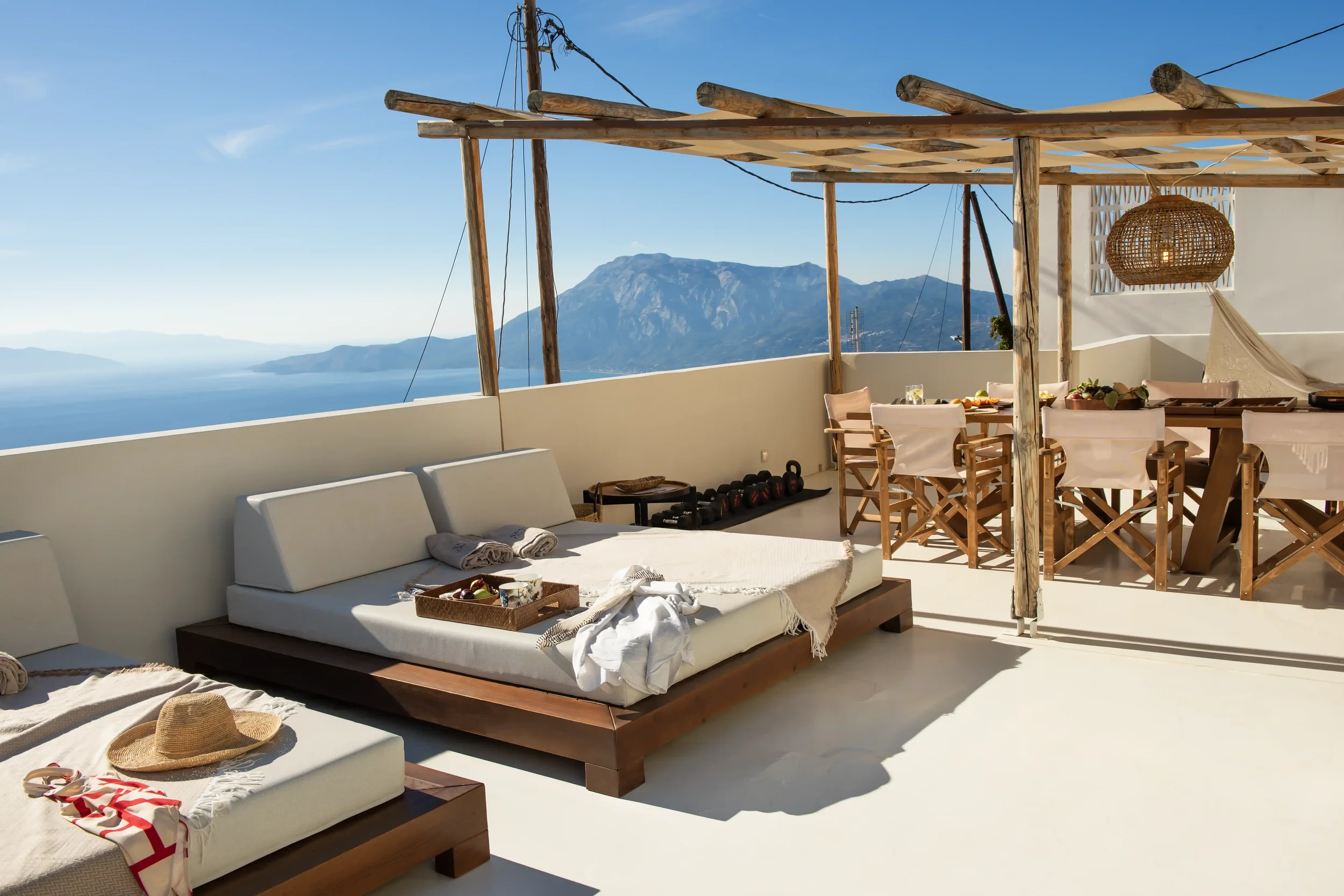 Sunlit outdoor terrace with white cushioned seating, wooden table and chairs, overlooking a mountain and ocean view, with a wooden pergola and minimalist decor.