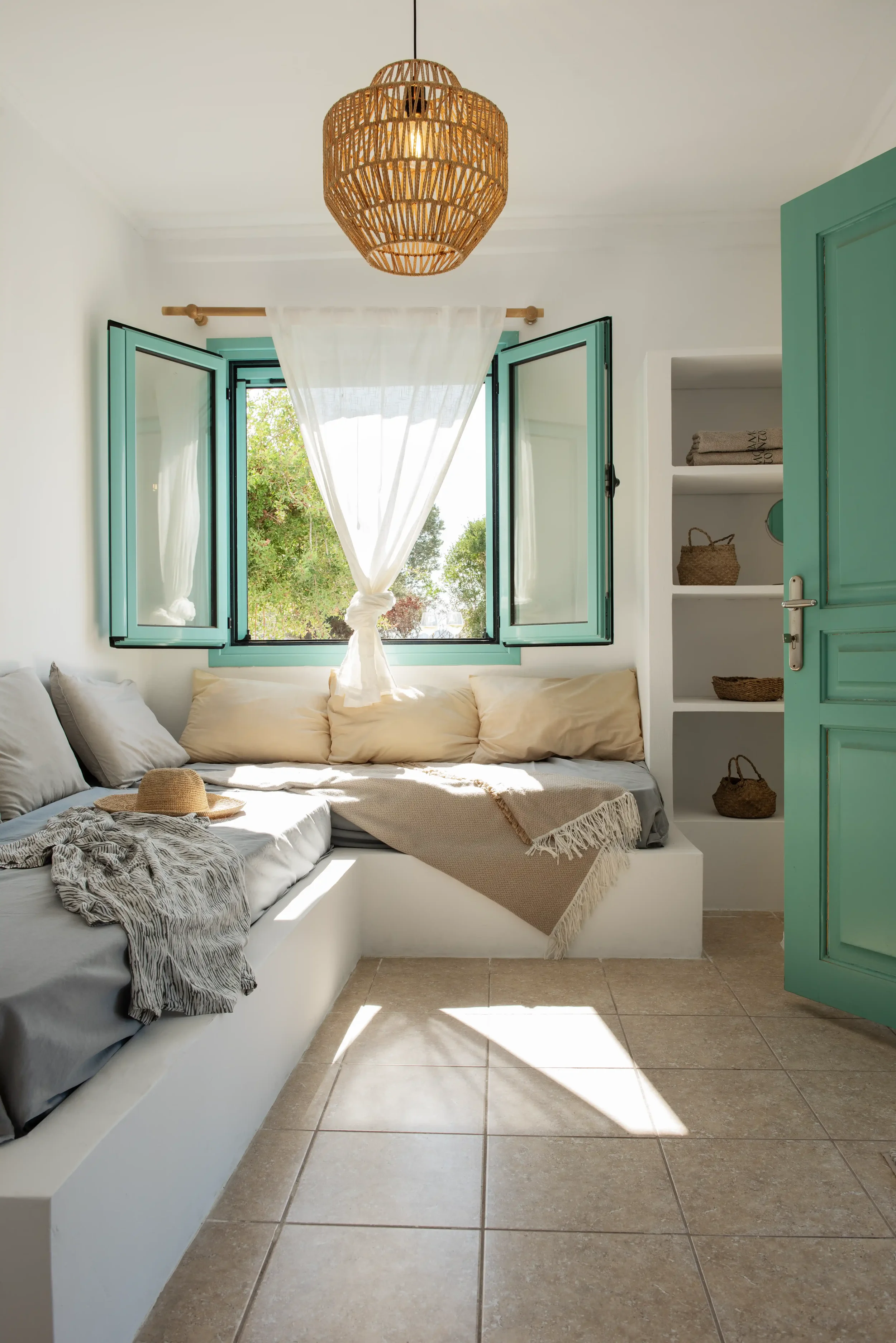 A cozy sunlit room with open turquoise window shutters, a white built-in sofa with beige and gray pillows, a straw hat and blanket on it, and a small white shelf with wicker baskets, all decorated in a bright, airy style.