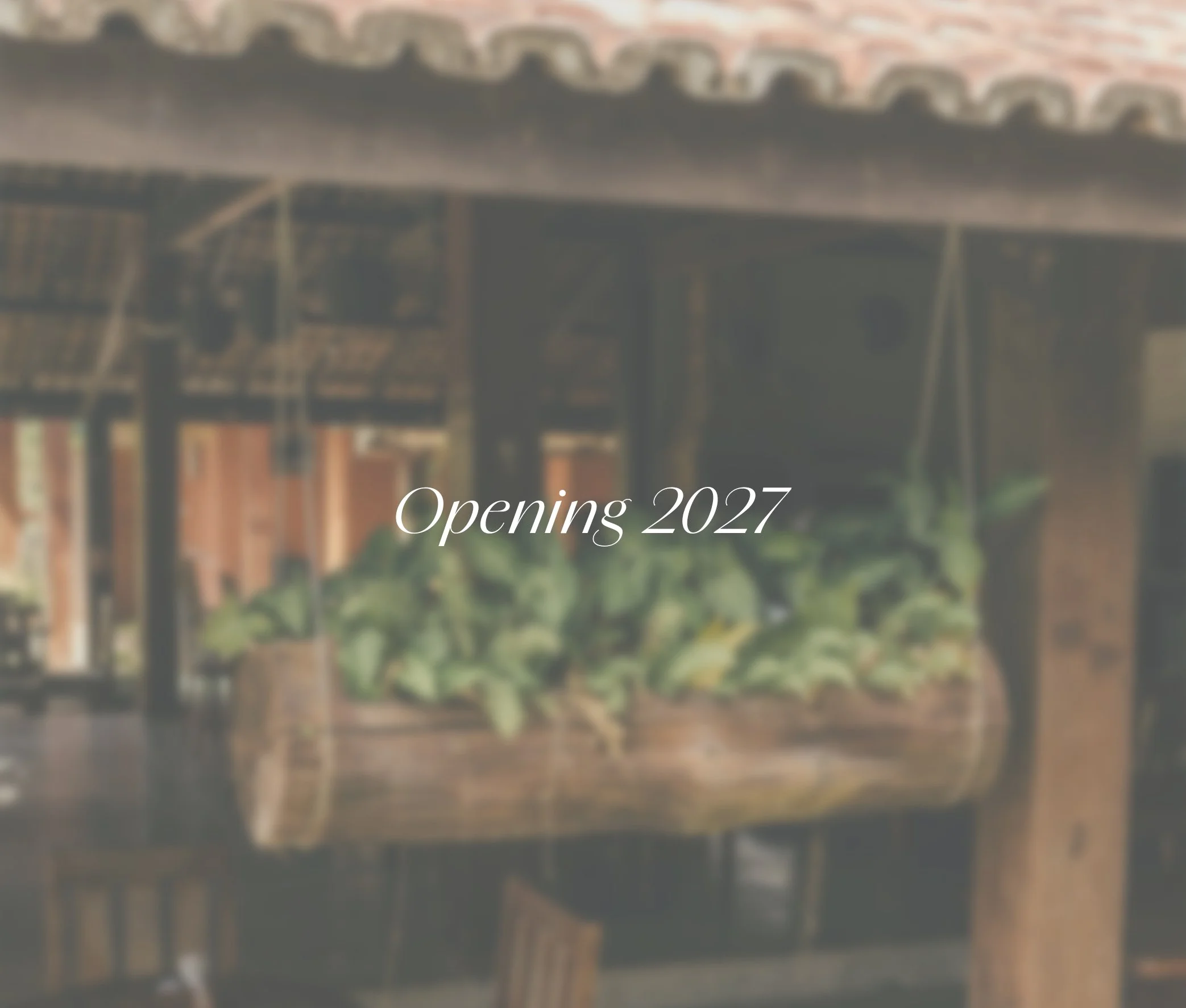 Blurred photo of a rustic, open-air structure with wooden beams and a tiled roof, displaying plants and a barrel, with text overlay that reads 'Opening 2027'.