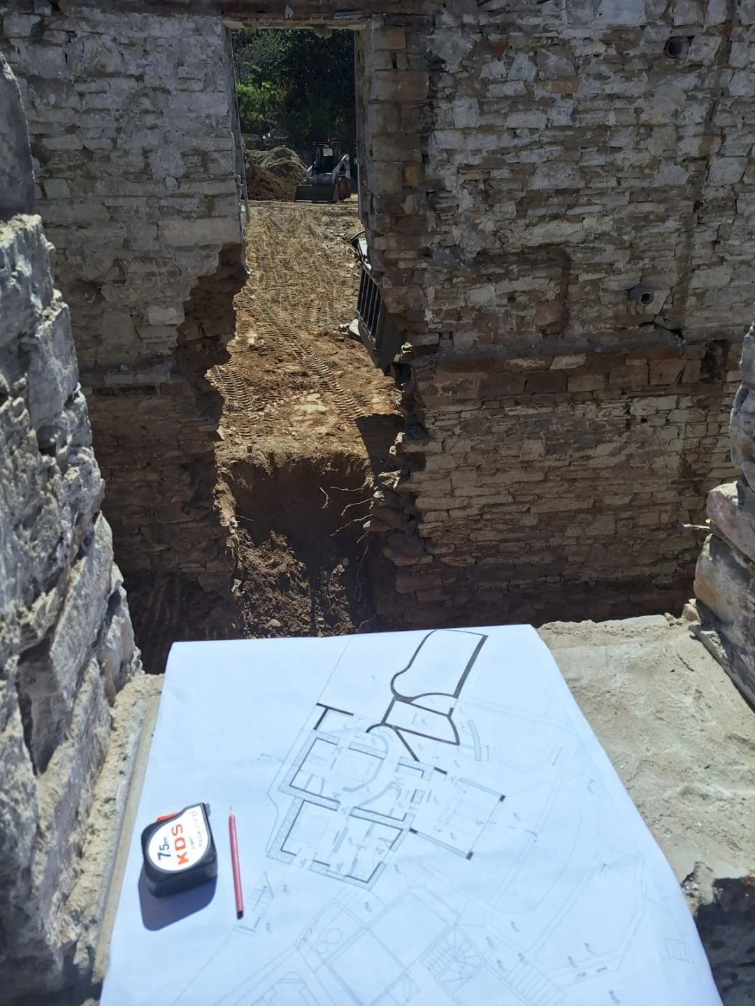 A construction site with a partially demolished brick wall, a blueprint on the ground, and construction equipment in the background.