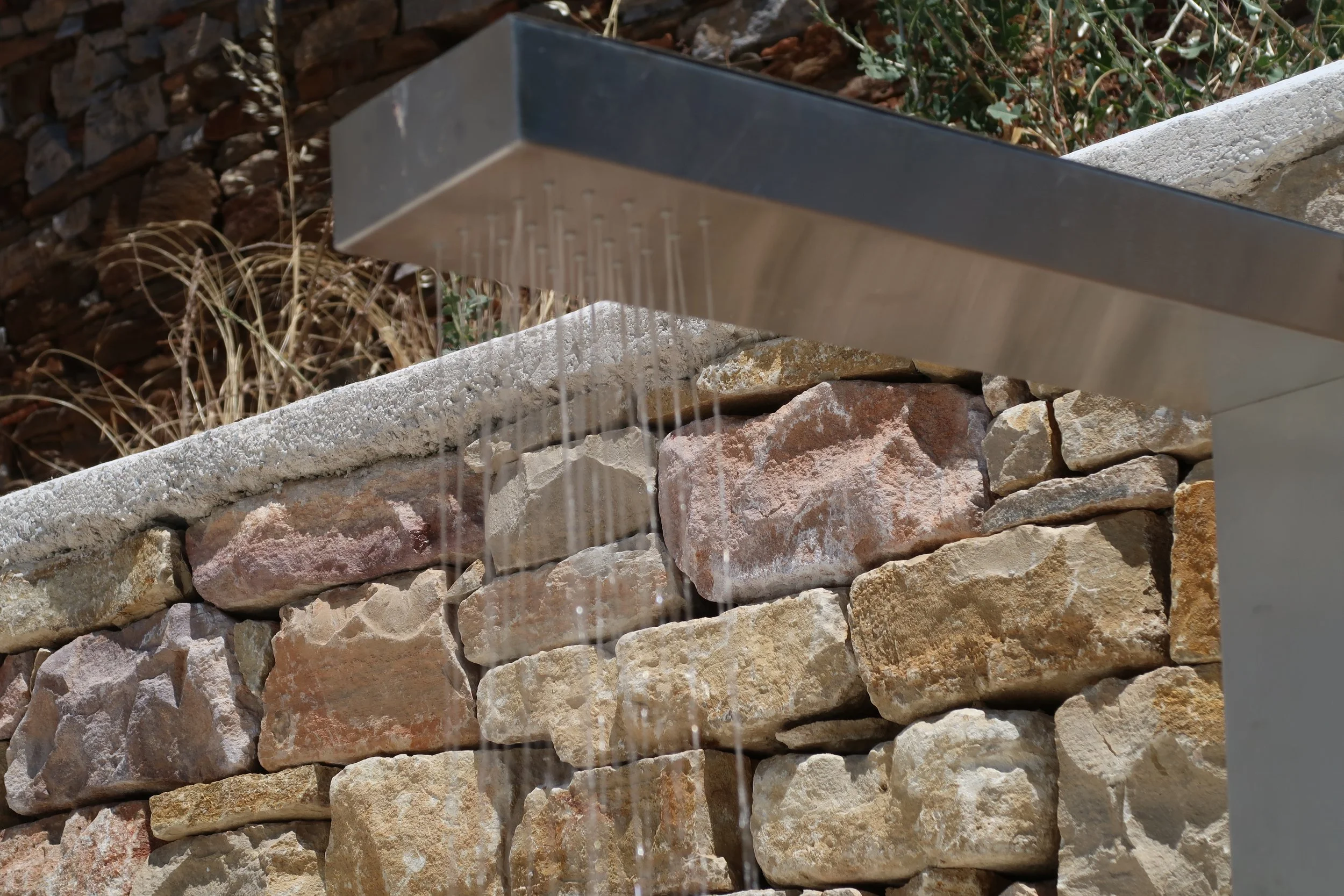 Water flows from a metallic water feature over a stone wall with various sizes and colors of rocks, set outdoors.