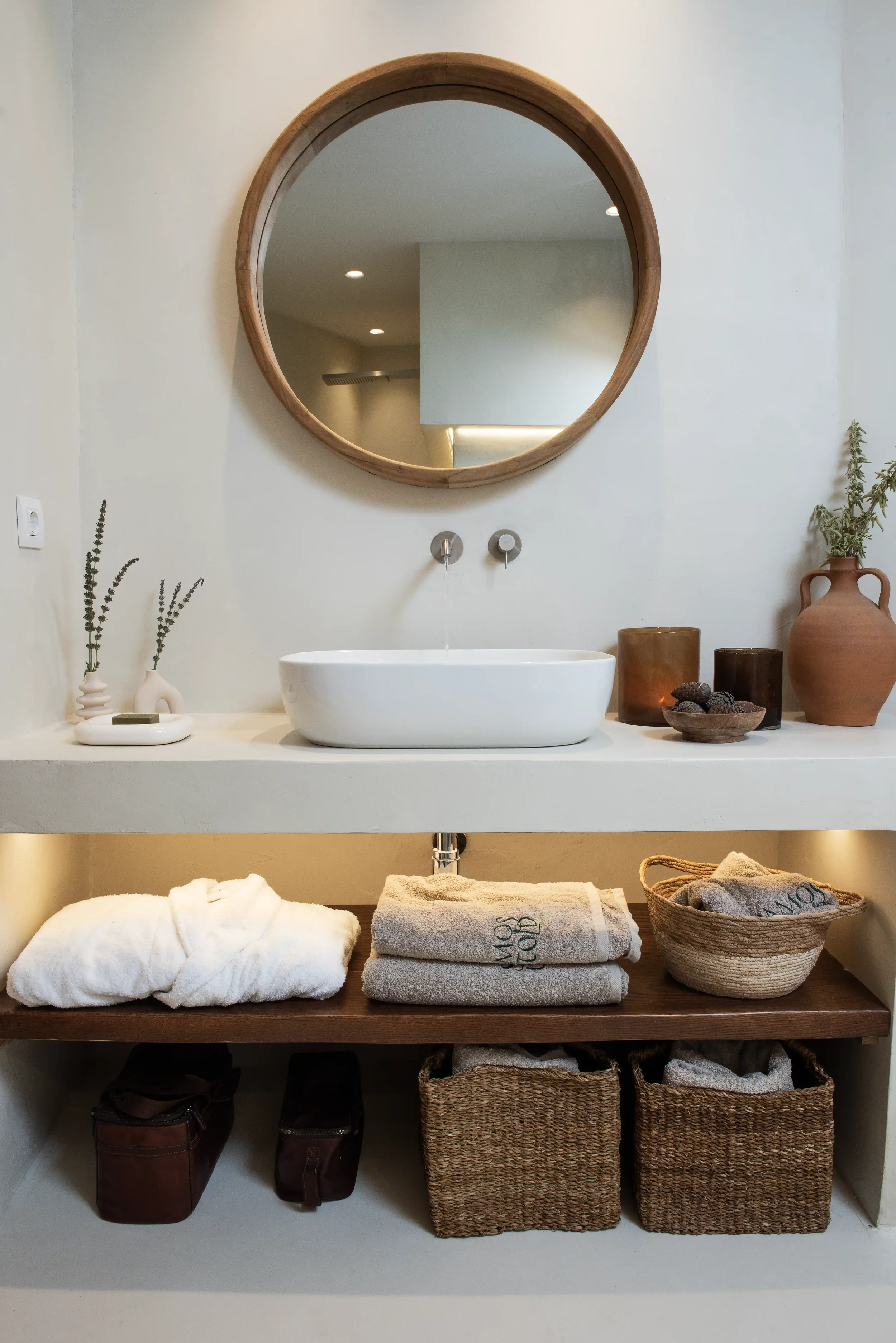 A bathroom vanity with a white vessel sink, a round wooden framed mirror, decorative vases and objects, and neatly folded towels and baskets underneath.