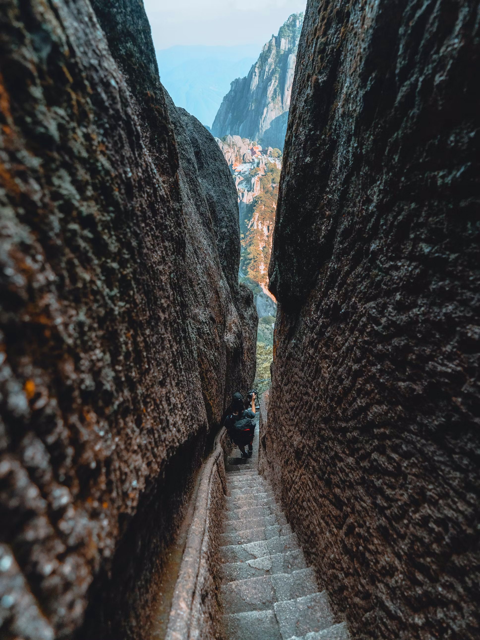 Person climbing a narrow stone staircase between large, textured rock walls with a scenic mountain village in the distance.