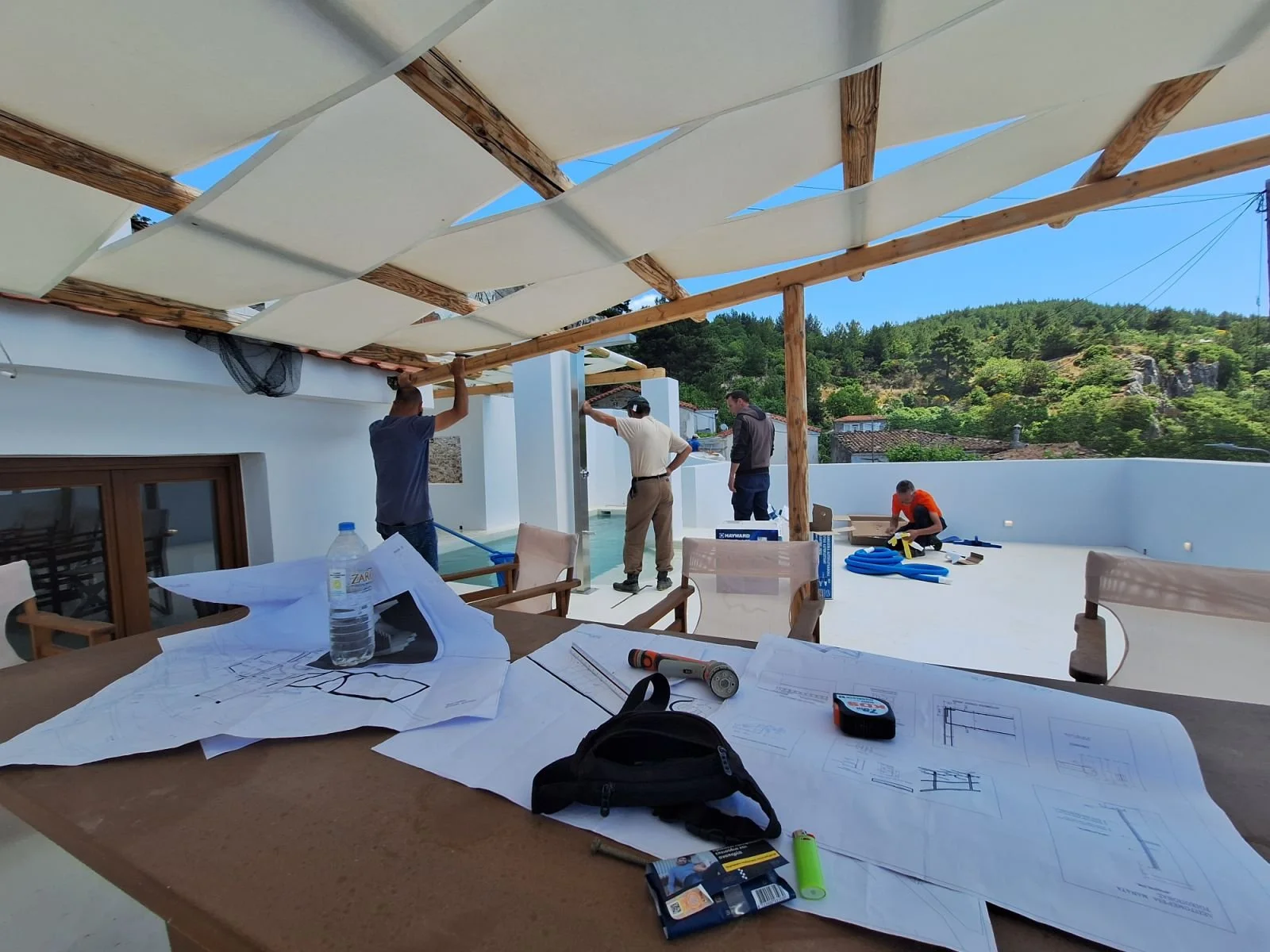 Construction workers installing a shaded roof on a terrace with tools, blueprints, and a water bottle on a table in the foreground.