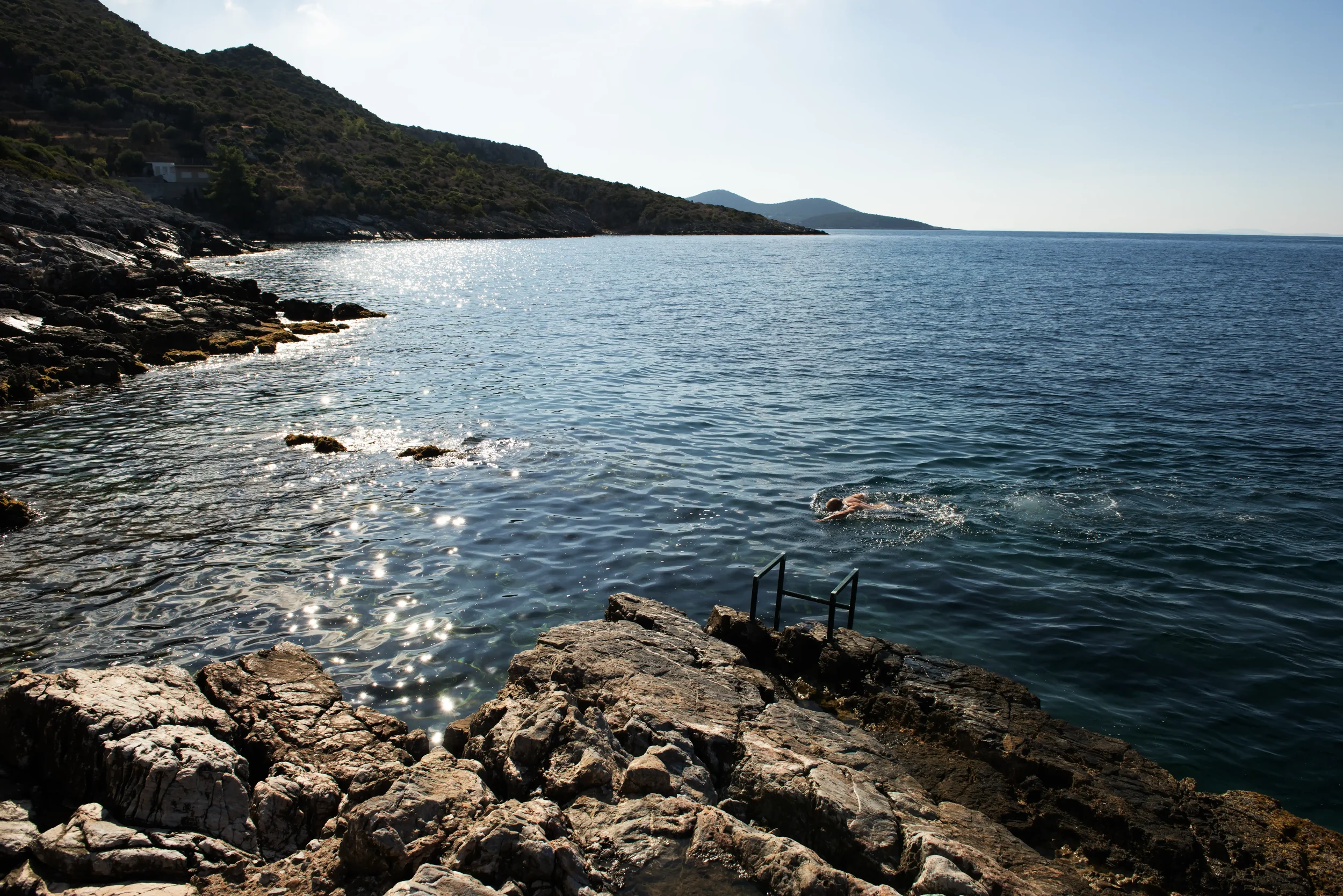 Person swimming in the ocean near rocky shoreline with hills in the background.
