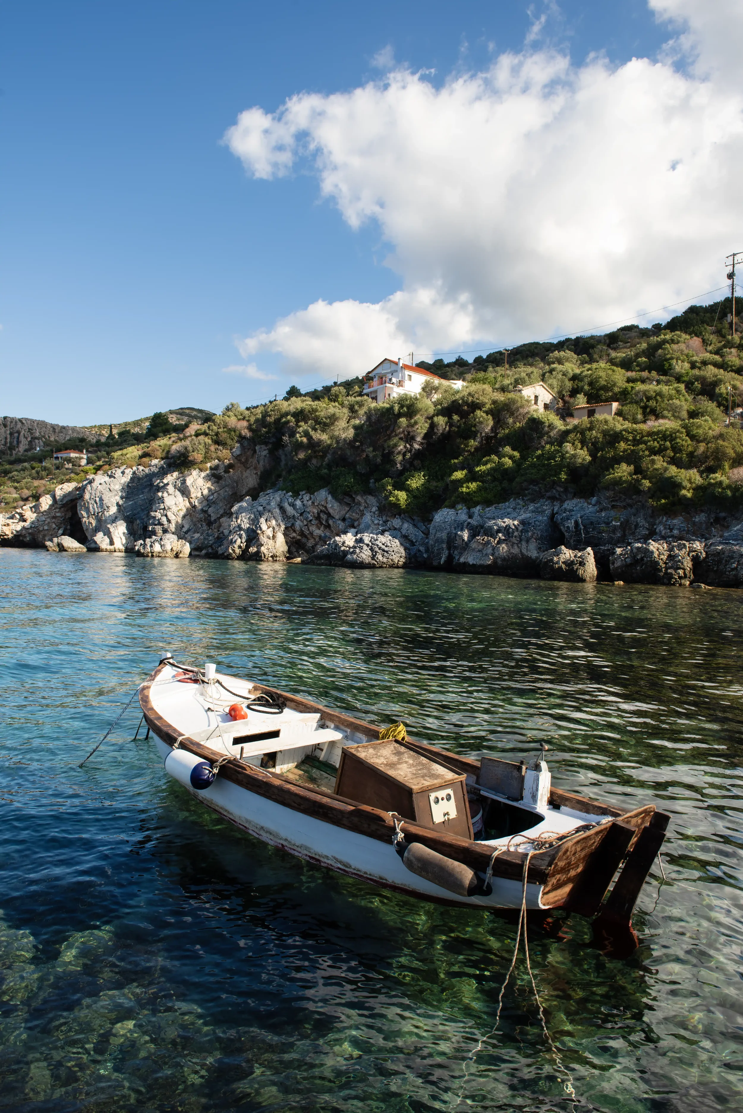 A small wooden boat floating on calm, clear water near the rocky shoreline with houses and greenery on the hillside in the background under a partly cloudy sky.