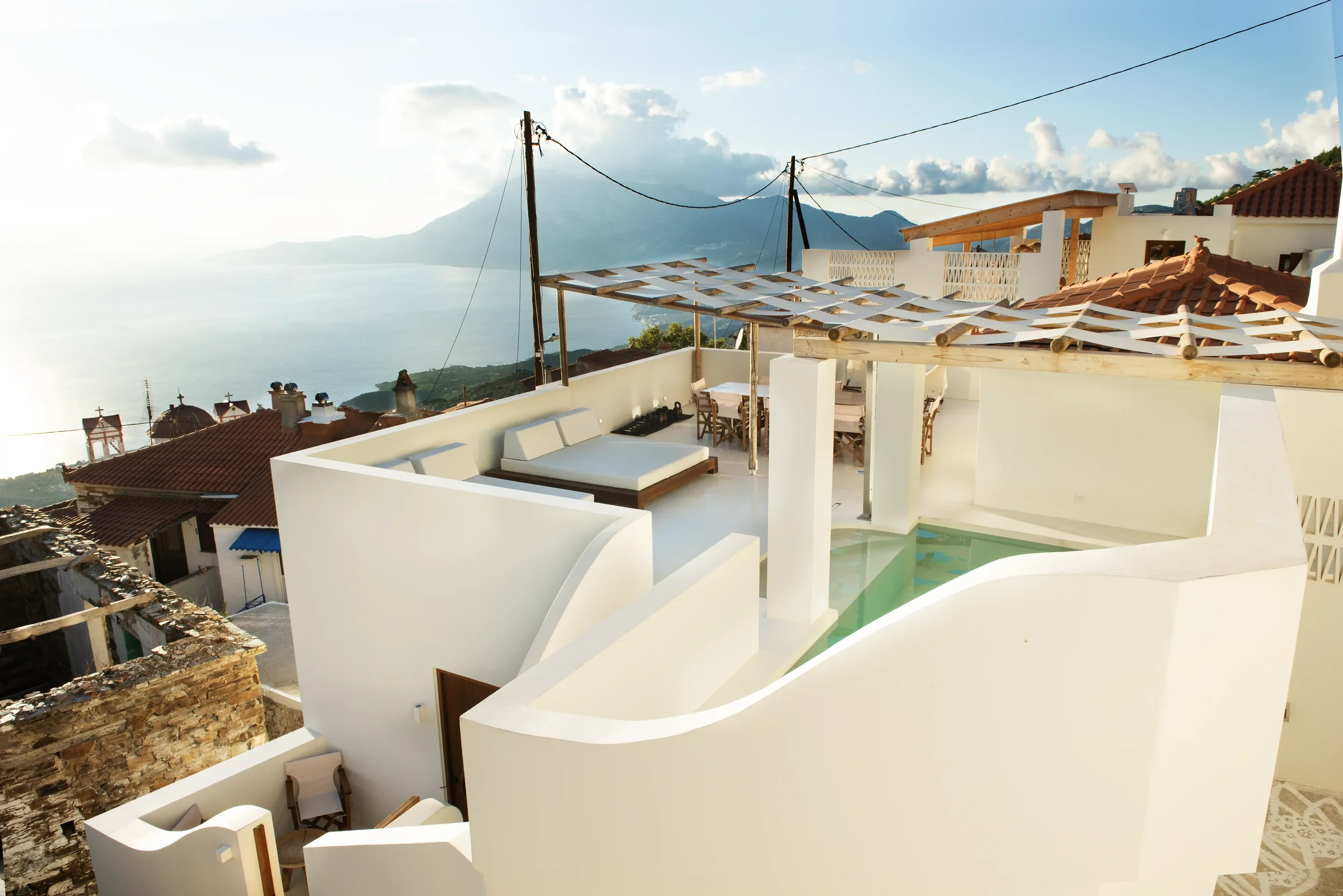 A rooftop terrace with white walls, outdoor seating, a small pool, and mountain views in the background during daytime.