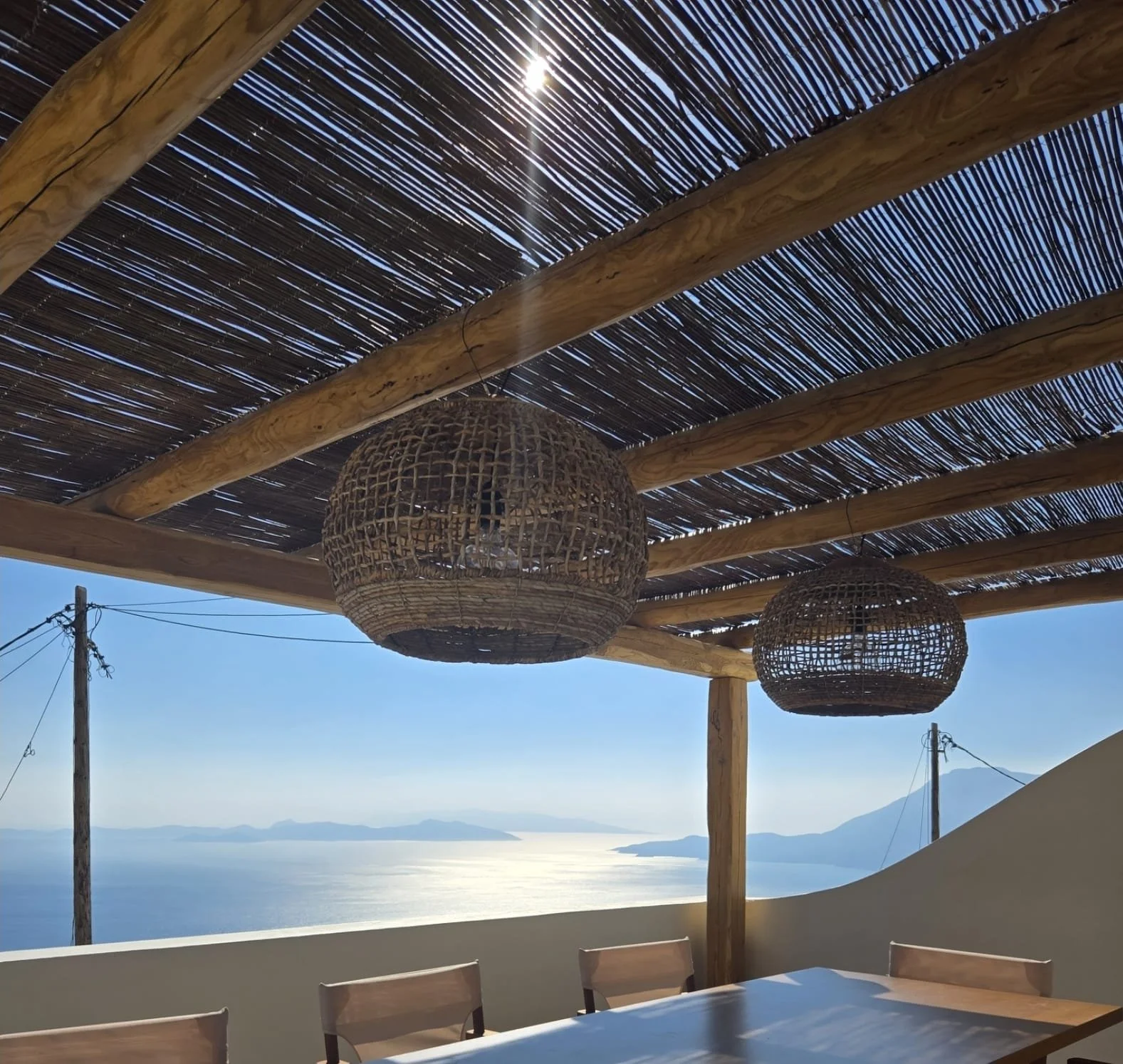 Outdoor dining area with a wooden table and chairs under a pergola with woven rattan lanterns, overlooking a body of water and distant islands or mountains.