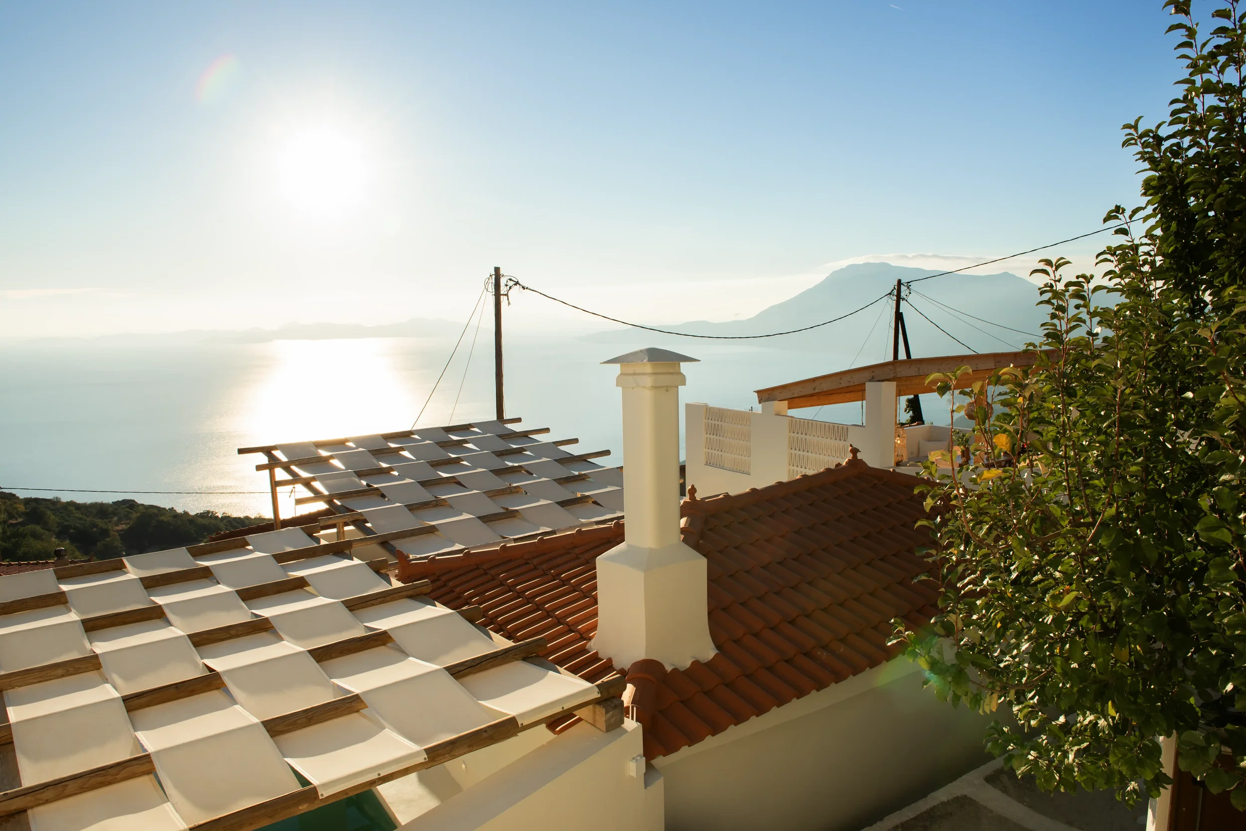 View of rooftops with red tiles and a chimney, overlooking the ocean with the sun shining brightly and mountains in the distance, and some trees in the foreground.