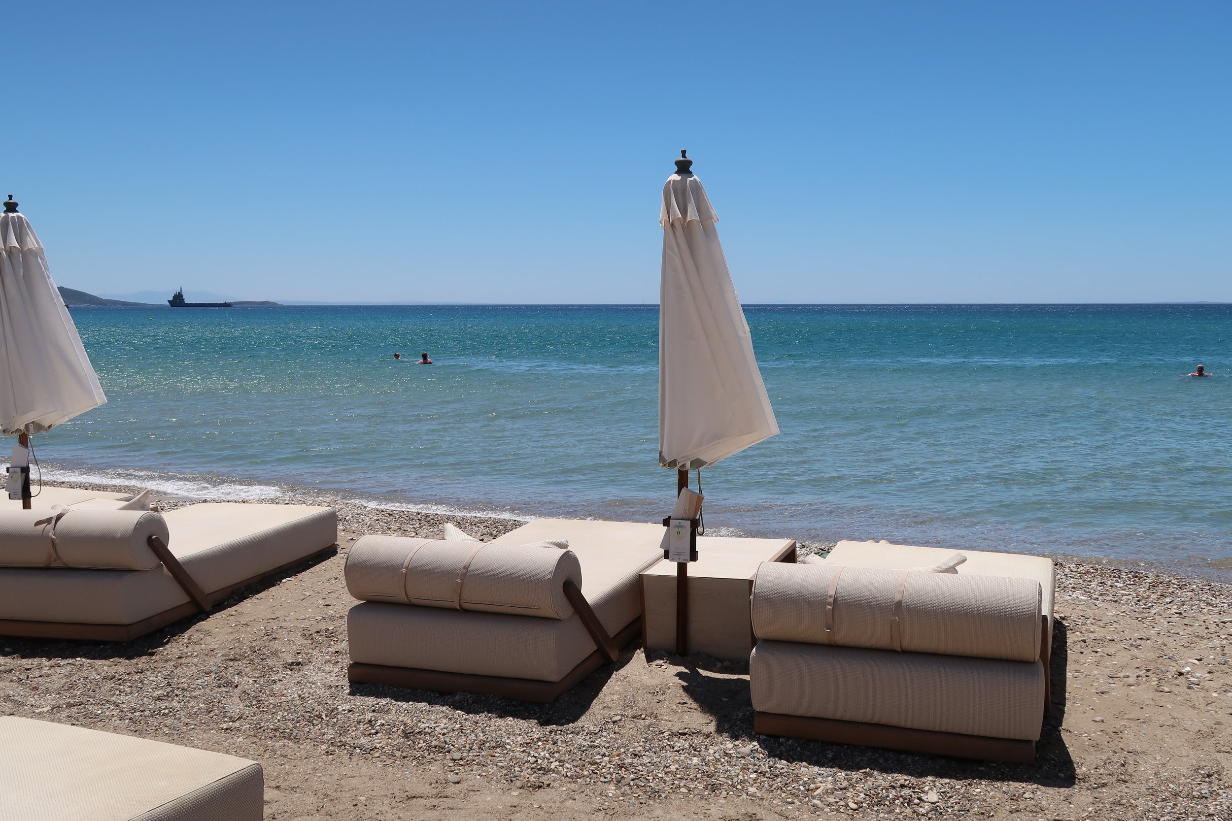Empty beach loungers and umbrellas facing the sea on a sunny day with a few people swimming in the ocean and a ship in the distance.