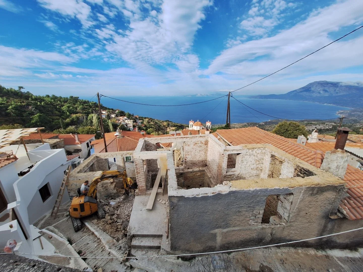 Construction site on a hillside overlooking a coastal town and the ocean, with a partly demolished building, a small excavator, red-tiled roofs, greenery, and a mountain in the background.