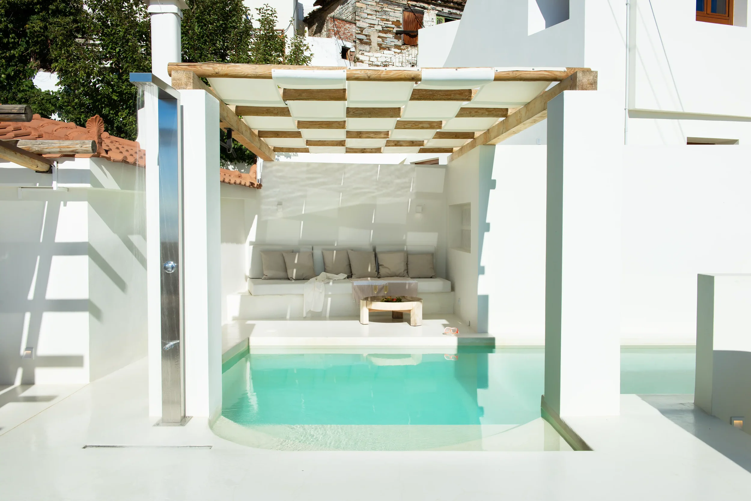 A small swimming pool in a modern outdoor space with a shaded sitting area featuring a white built-in couch and beige cushions, surrounded by white walls and a wooden pergola.
