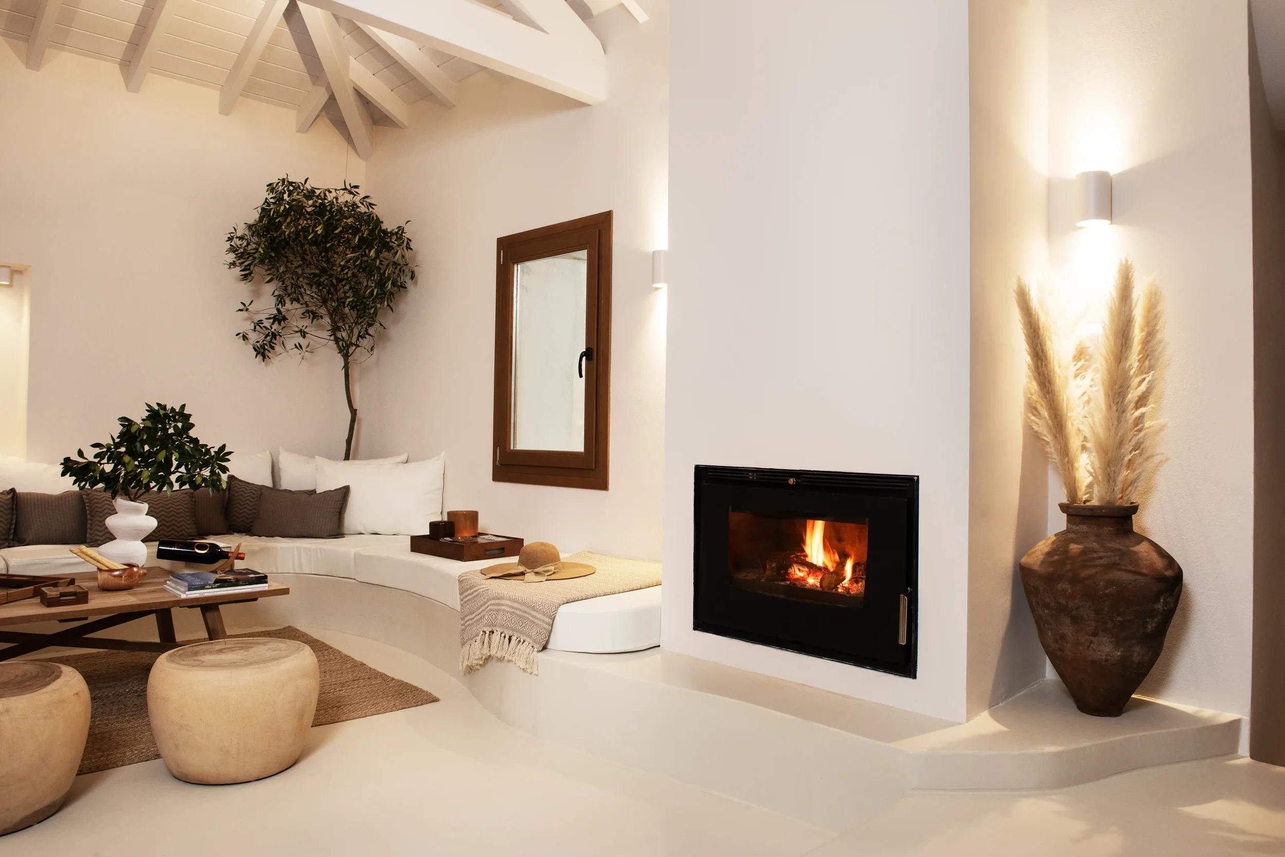 Modern living room with white walls, wooden ceiling beams, a built-in white sofa with gray and white pillows, a wooden coffee table, a fireplace with a lit fire, a large decorative vase with pampas grass, and a window with wooden frame.