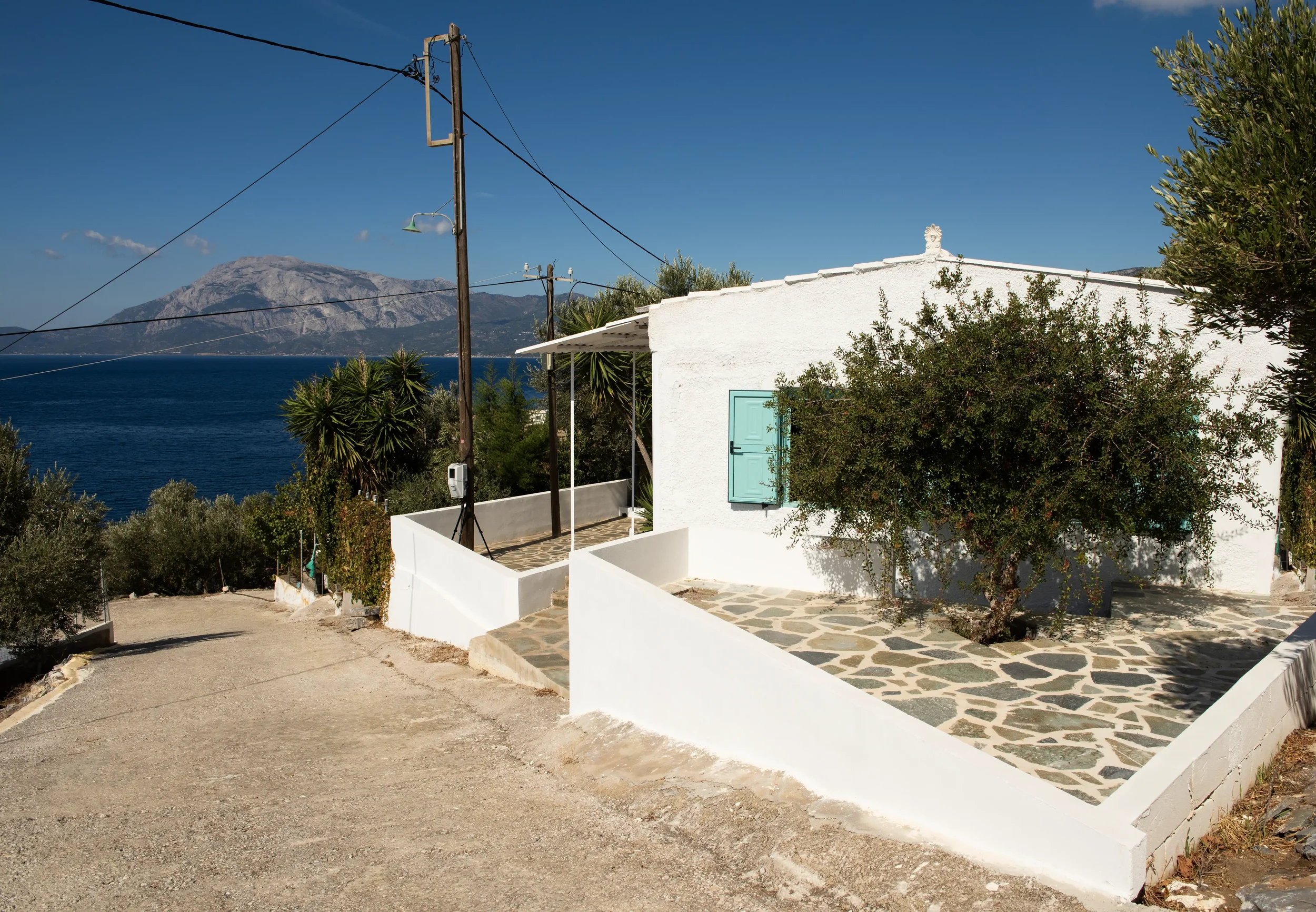 A white Mediterranean-style house with a light blue window shutter, surrounded by greenery, overlooking a body of water with a mountain in the background under a clear blue sky.
