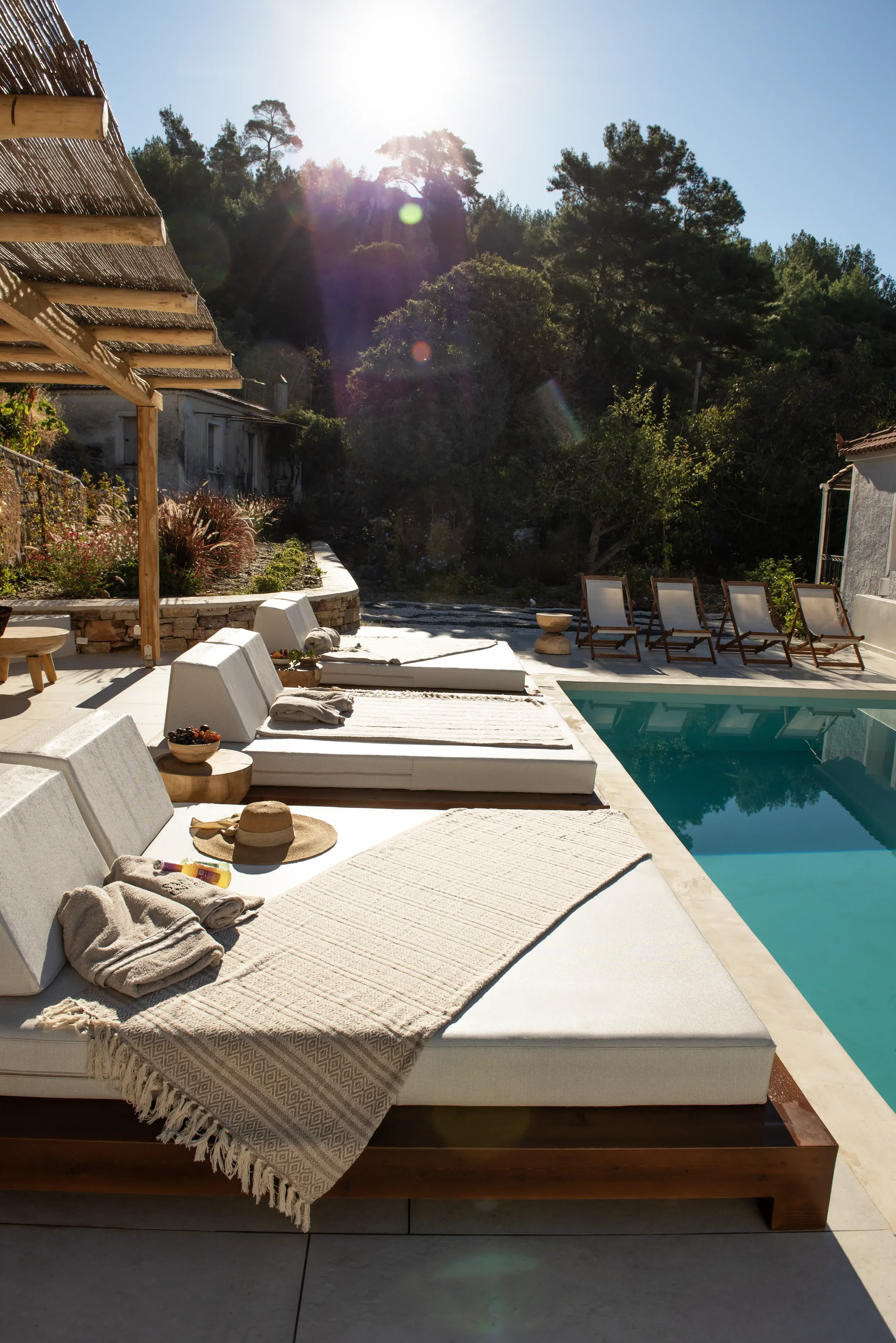 Sunlit outdoor poolside lounge area with cushioned sunbeds, towels, a straw hat, and a bowl of berries, set against a backdrop of trees and hillside.