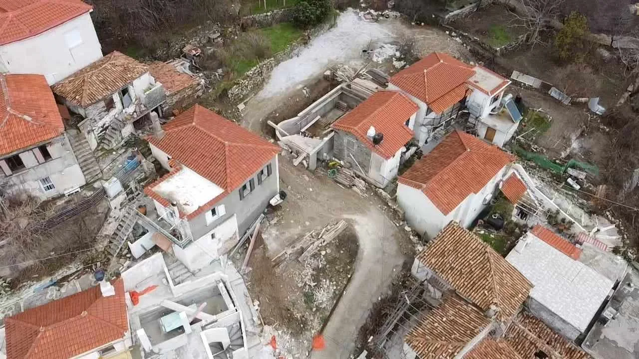 Aerial view of a neighborhood with multiple houses featuring red-tiled roofs, some under construction or repair, with a muddy, unpaved street winding through.