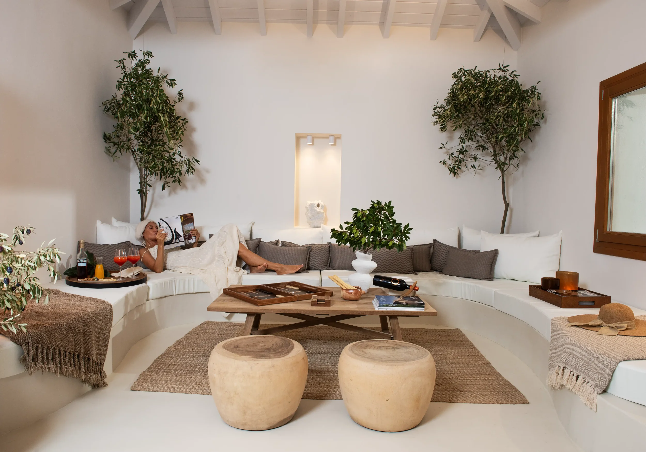 Modern, minimalist living room with a white curved sectional sofa, throw pillows, a wooden coffee table, and two rounded stools. A woman is relaxing on the sofa reading a magazine, with drinks and snacks on a tray nearby. The room has large green pot