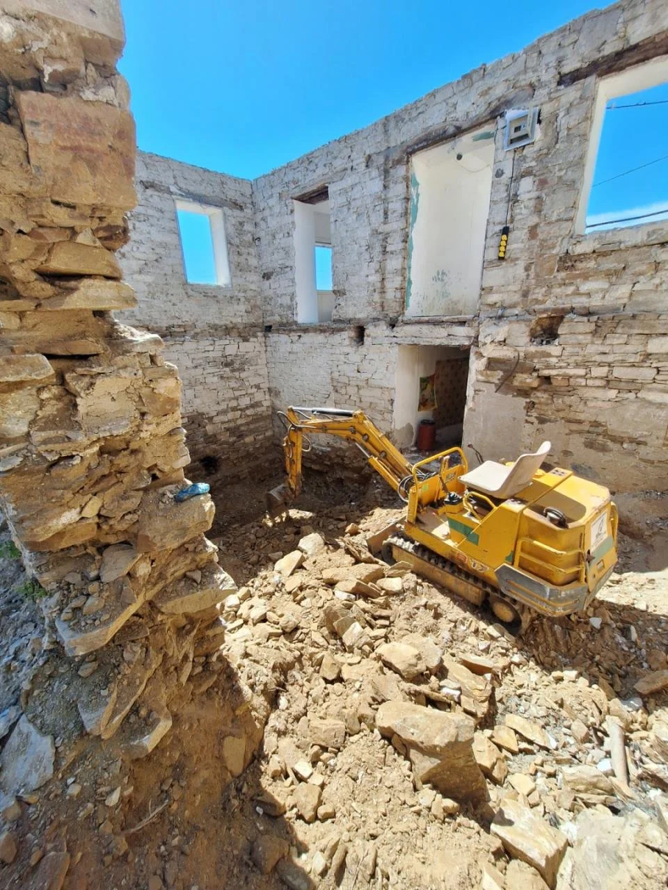 Construction site inside a building with exposed brick walls and large windows, featuring a small yellow excavator removing dirt and debris.