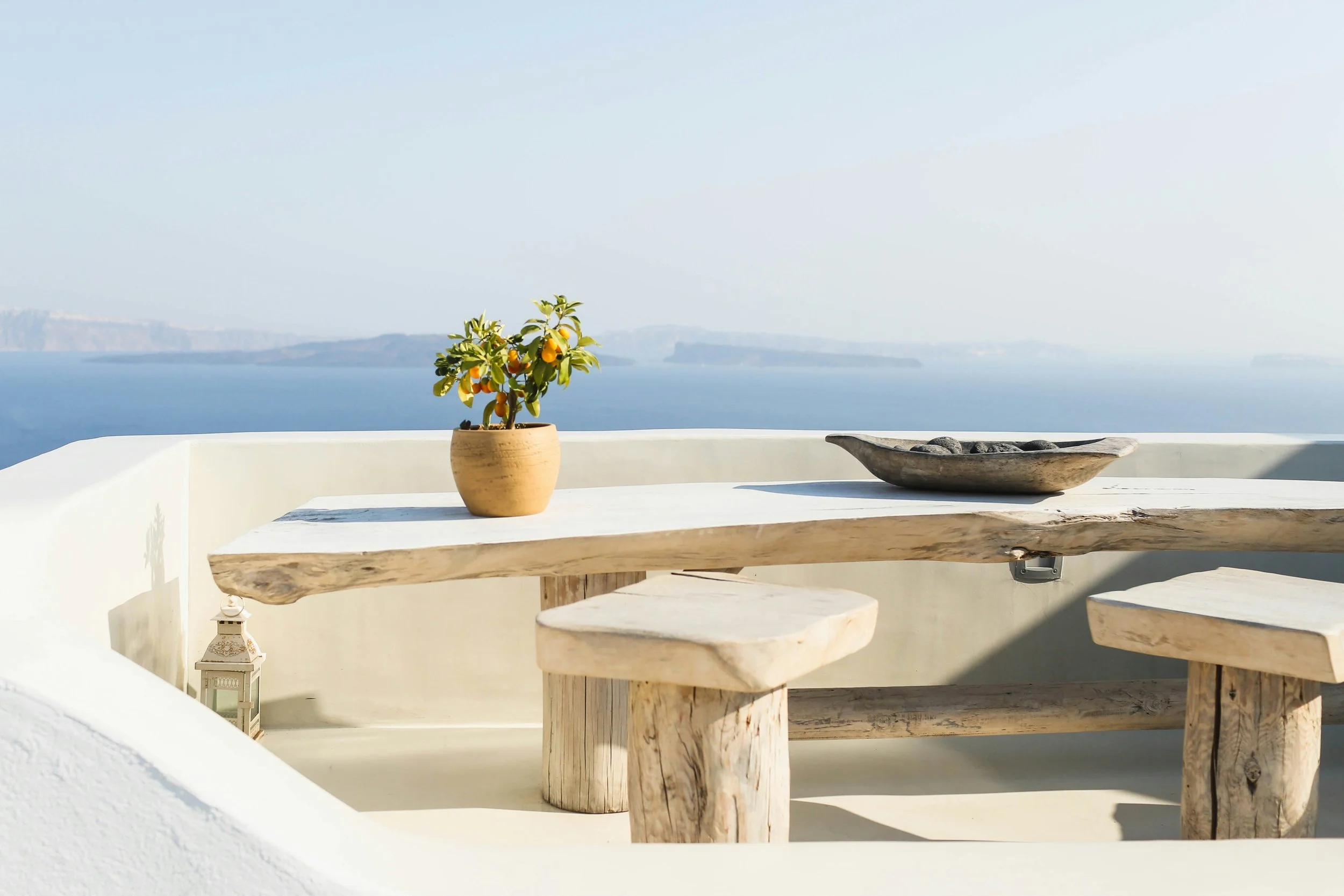 A rustic wooden table and benches on a white terrace overlooking the ocean, with a potted orange tree and a black tray with stones, interior of Samos Untold
