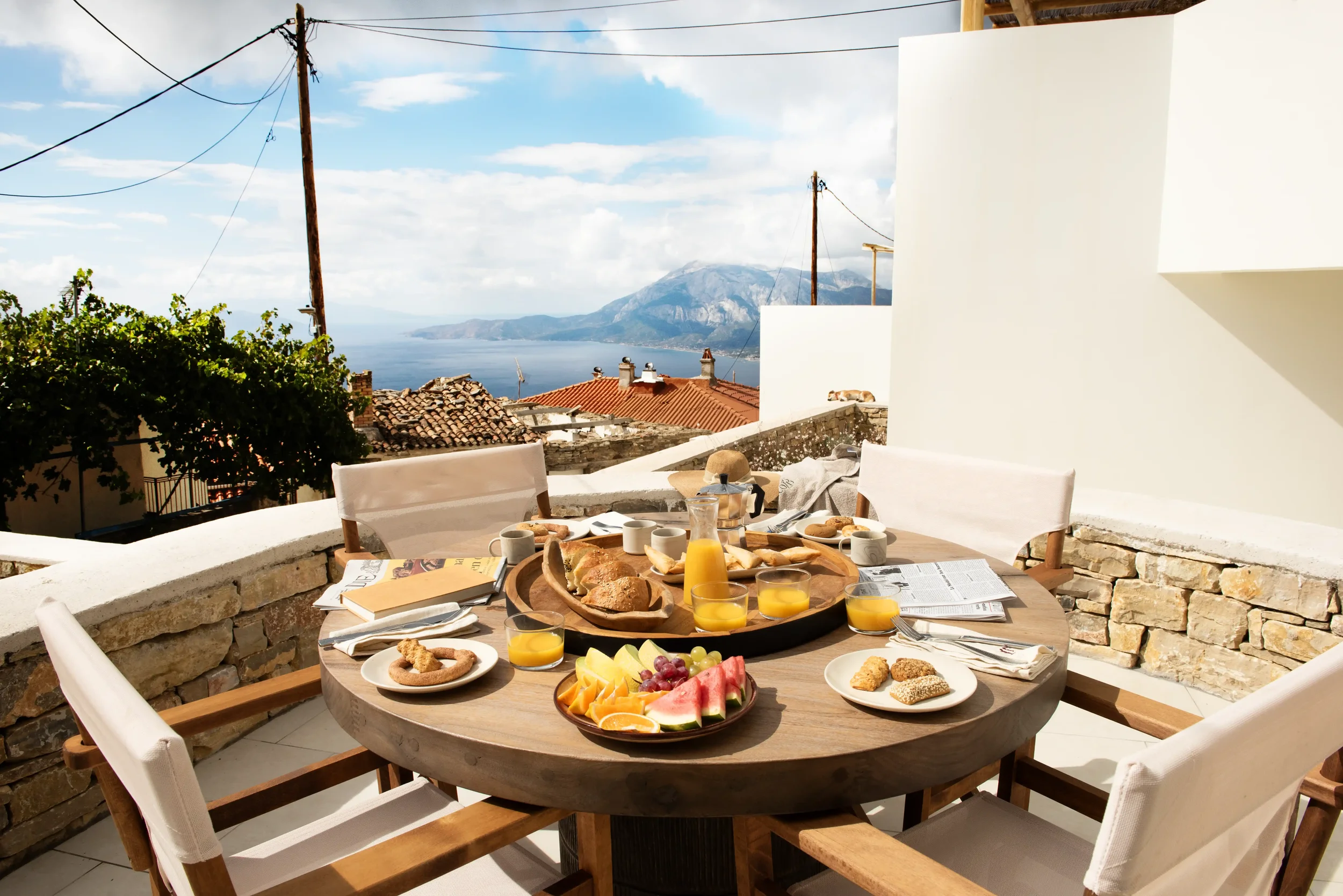 Outdoor breakfast table with assorted food and drinks, overlooking rooftops, the sea, and mountains under a partly cloudy sky.