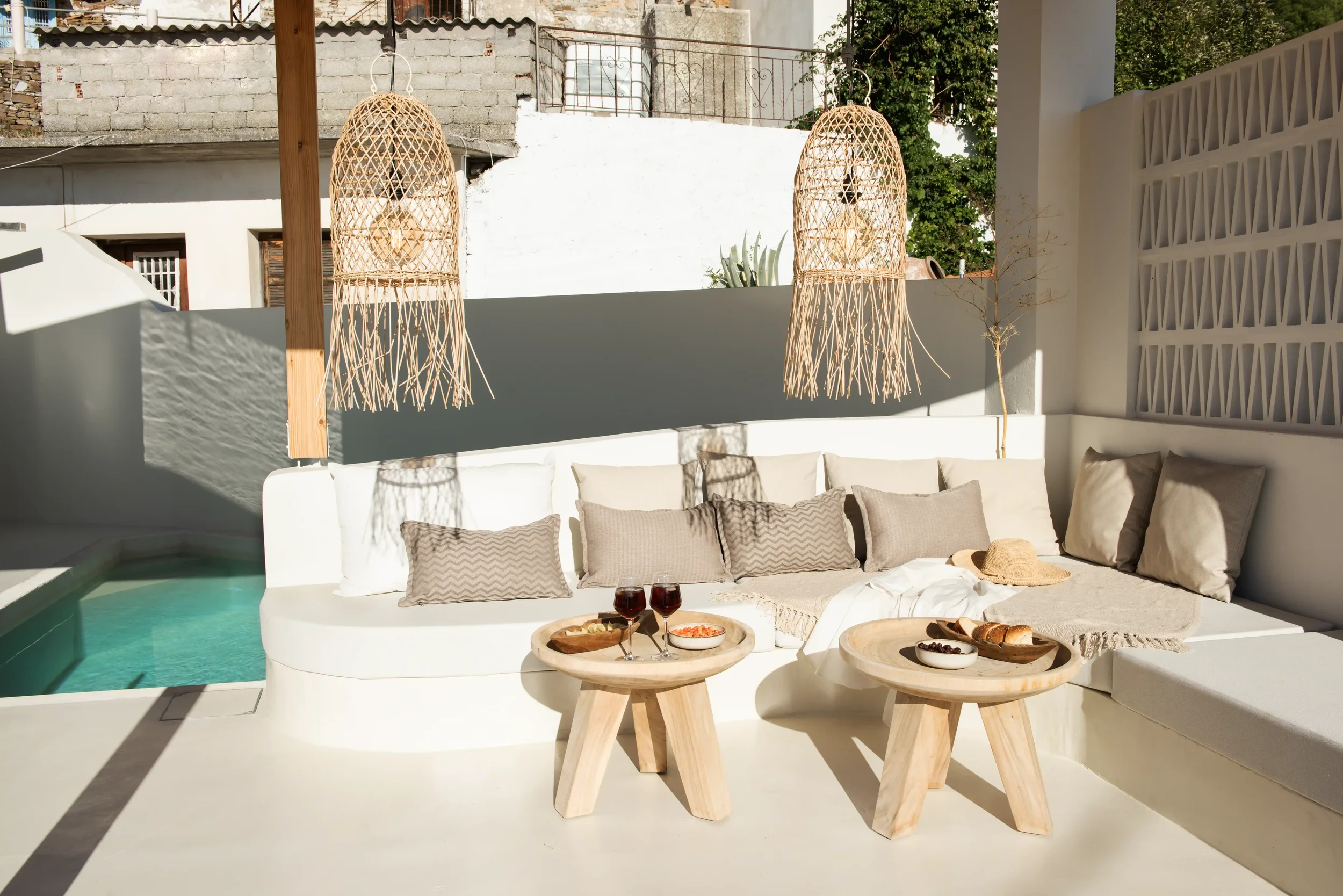 Outdoor sitting area with white cushioned seating, beige pillows, two wooden tables with snacks and drinks, two hanging wicker lights, and sunlight casting shadows.