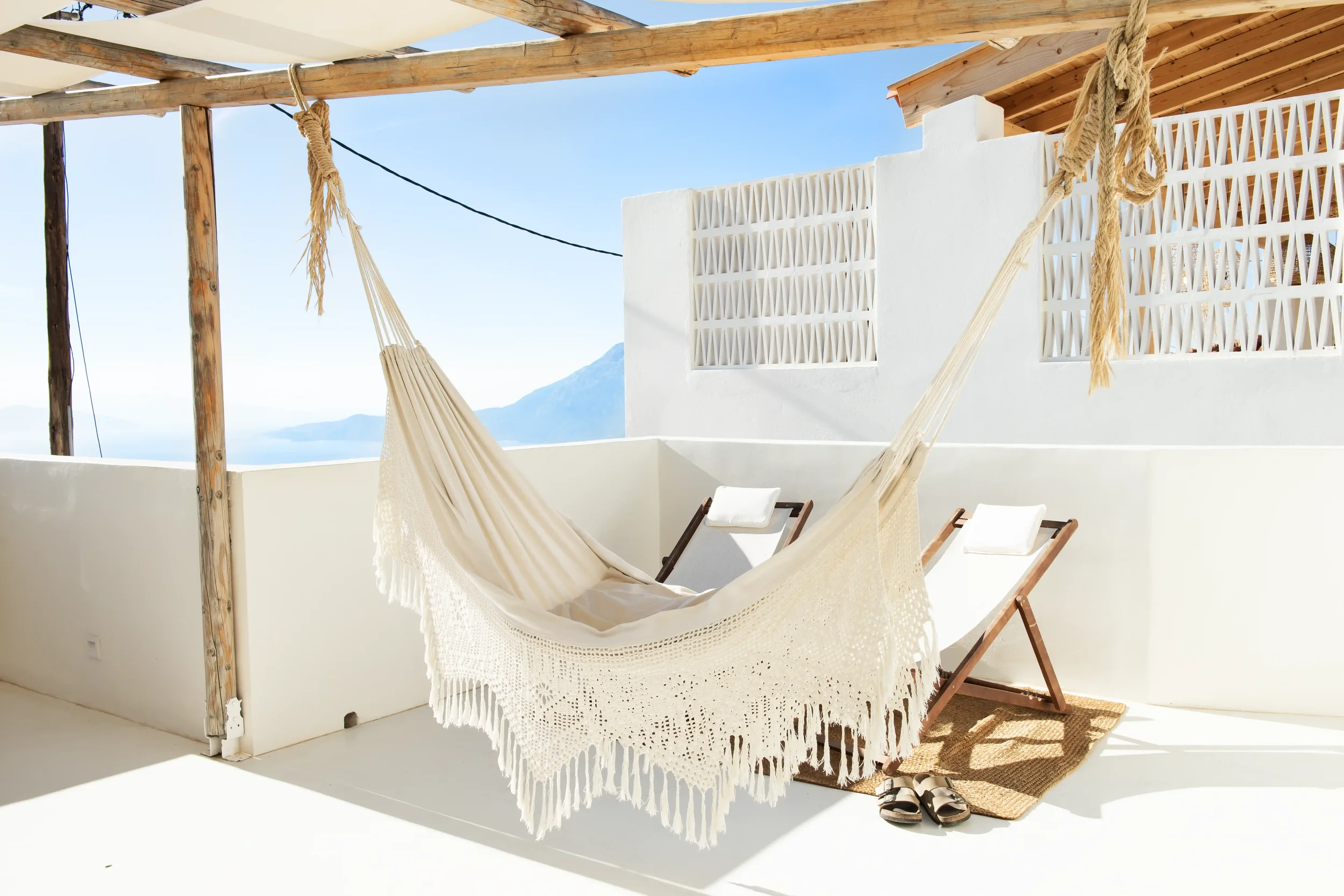 A balcony with a hammock and two deck chairs, with a scenic view of mountains and water in the background, under a wooden pergola.