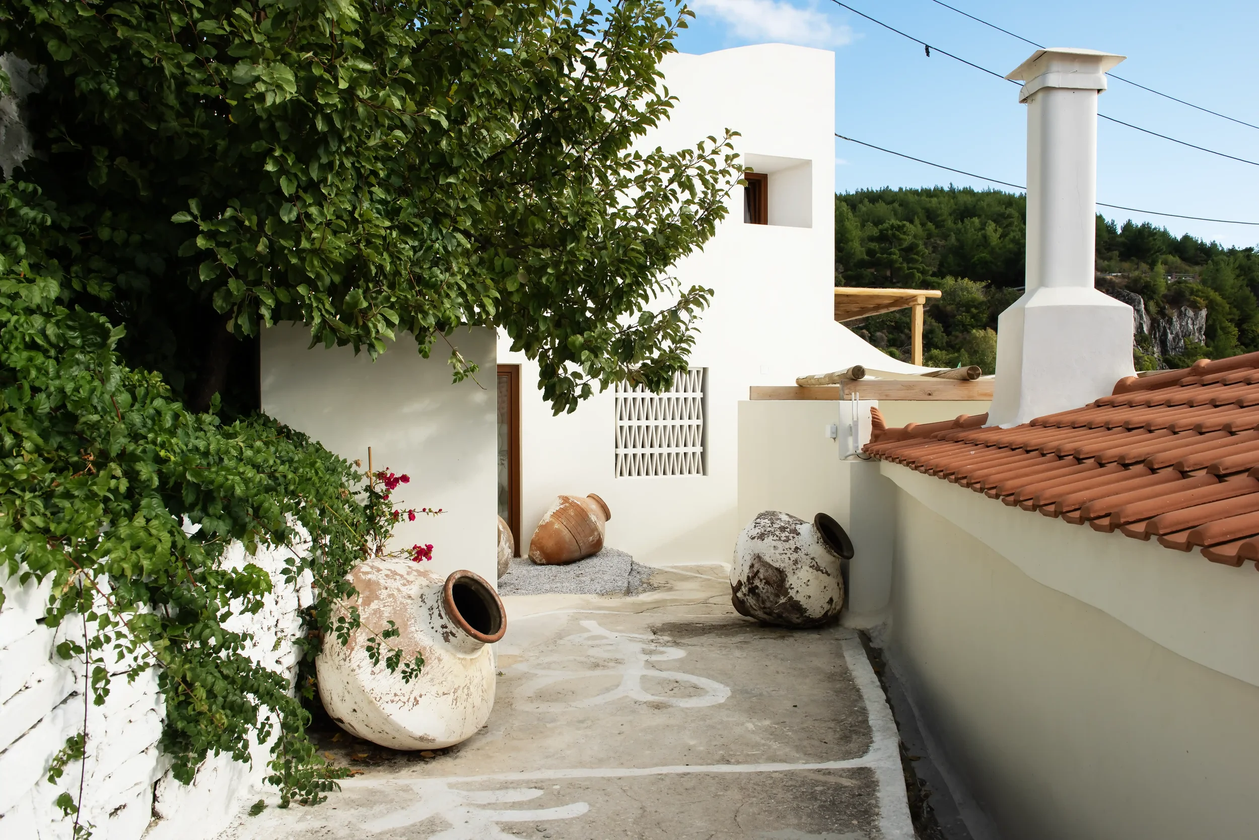 A Mediterranean-style terrace with white walls, large clay pots, a large tree, and red tiled roof, overlooking green hills.