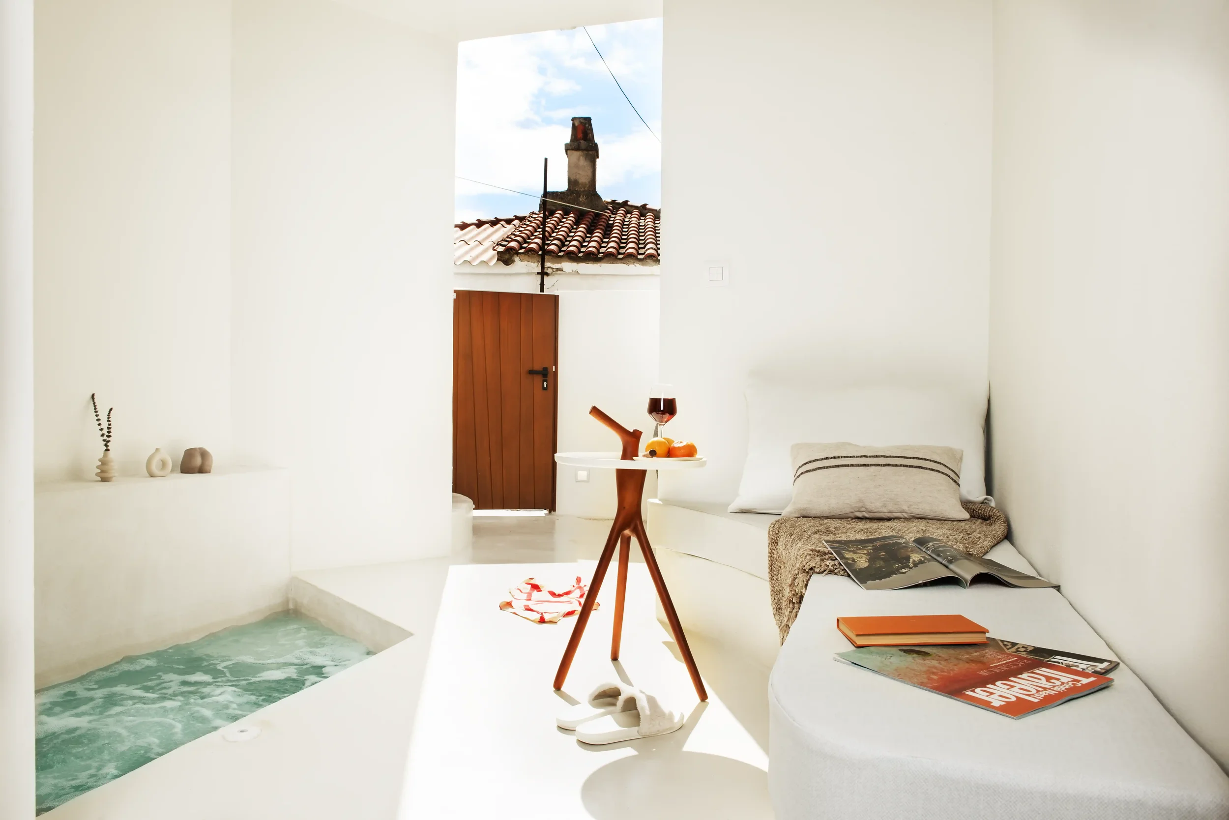 A cozy indoor space with a built-in white lounge and a small pool nearby, decorated with magazines, books, and a table with a glass of wine and a plate of fruit, overlooking a tiled roof and chimney outside.