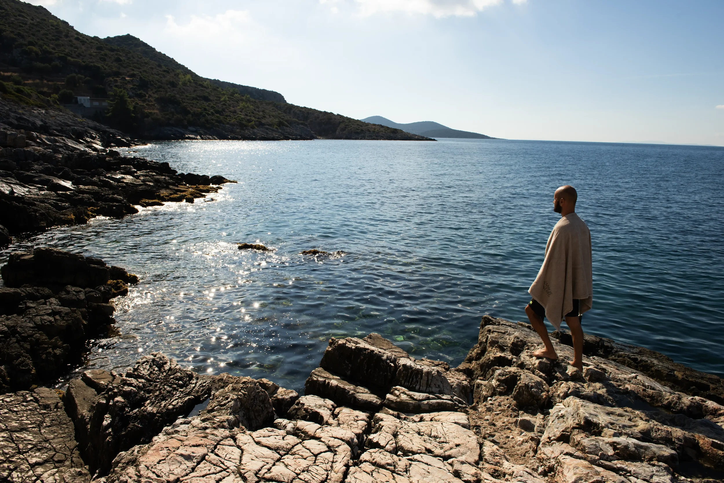 A man with a shaved head and beard standing on rocks by the water's edge, wrapped in a beige towel, overlooking a calm sea with hills in the background on a sunny day.
