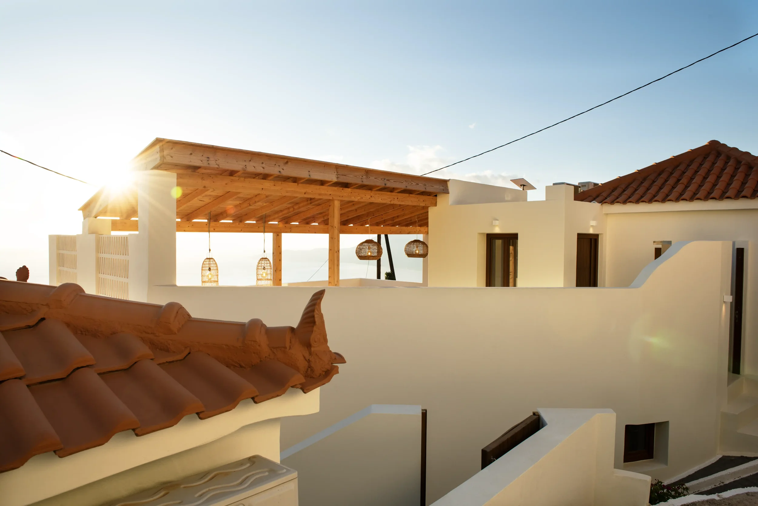 View of a white Mediterranean-style building with terracotta roof tiles, a wooden pergola, and hanging wicker light fixtures, during sunset or sunrise.