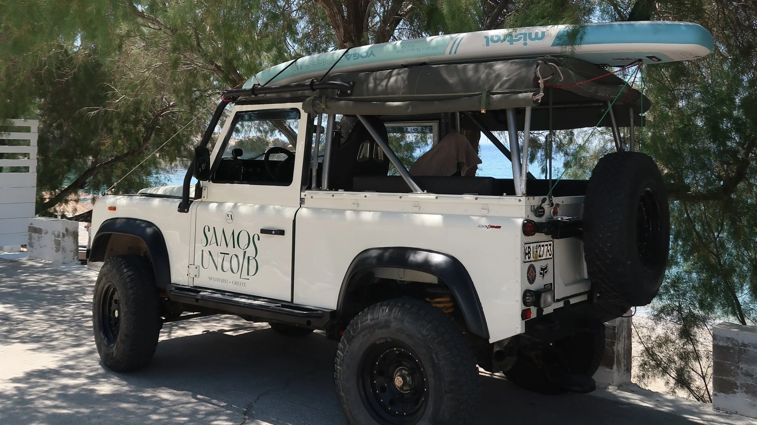 White off-road  Samos Untold vehicle with a roof rack holding a kayak and a paddle board, parked on a sandy area near trees with a view of the water in the background.