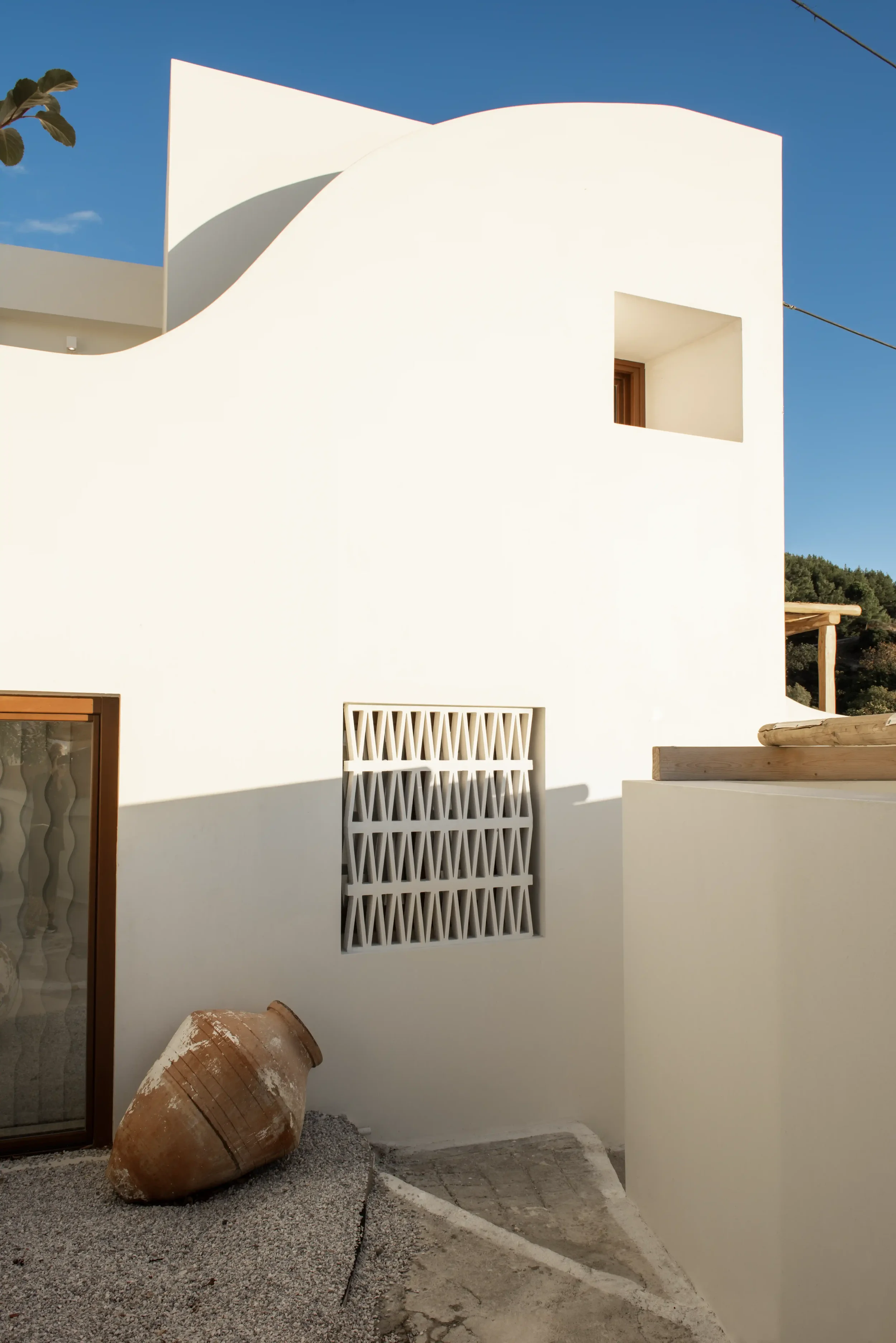 White modern villa with rounded and straight walls, wooden window frames, decorative grille, a large clay urn, and gravel ground under a blue sky.