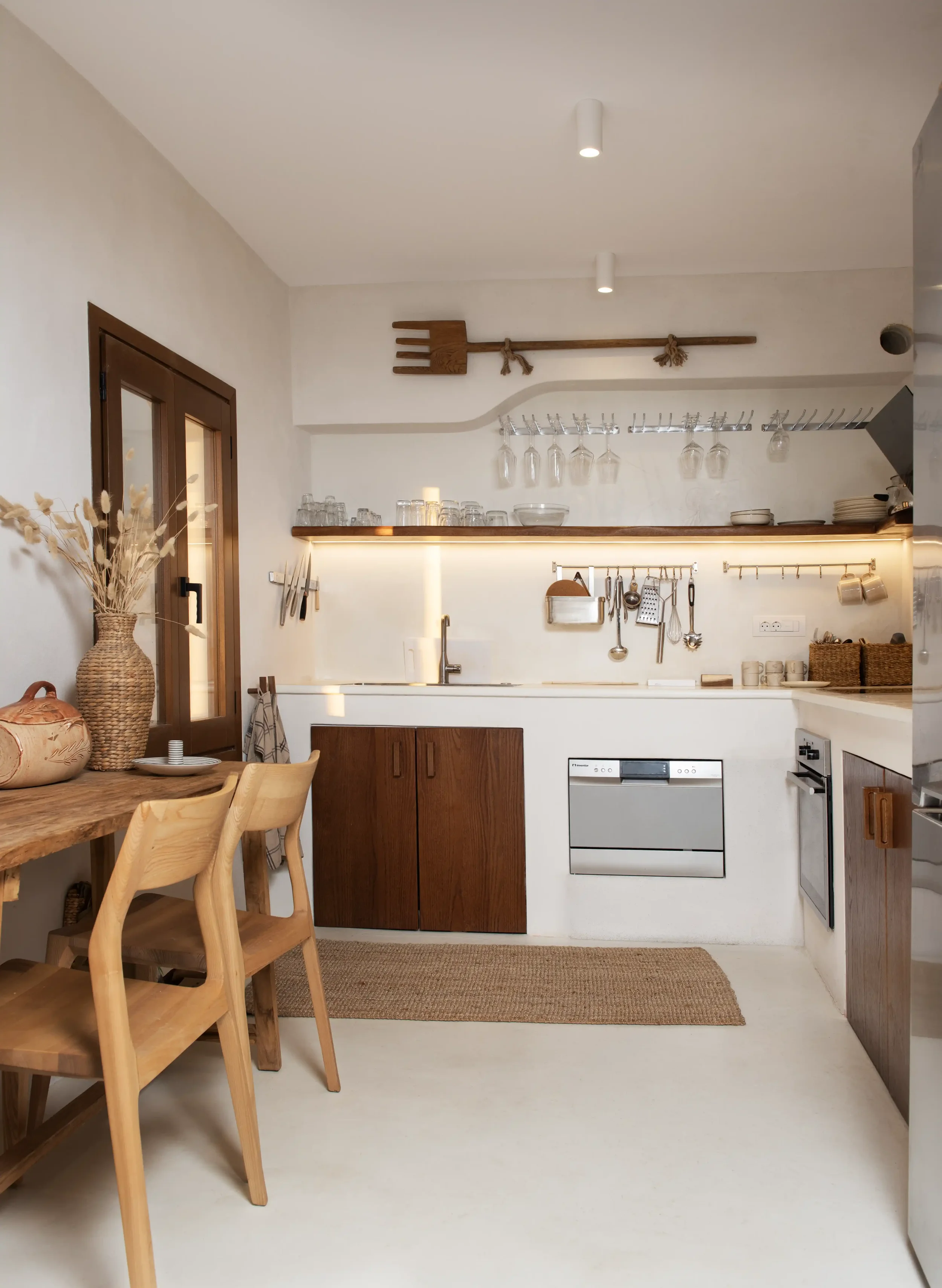 Minimalist kitchen with white walls, wooden accents, hanging glasses, and a dining table with wooden chairs.