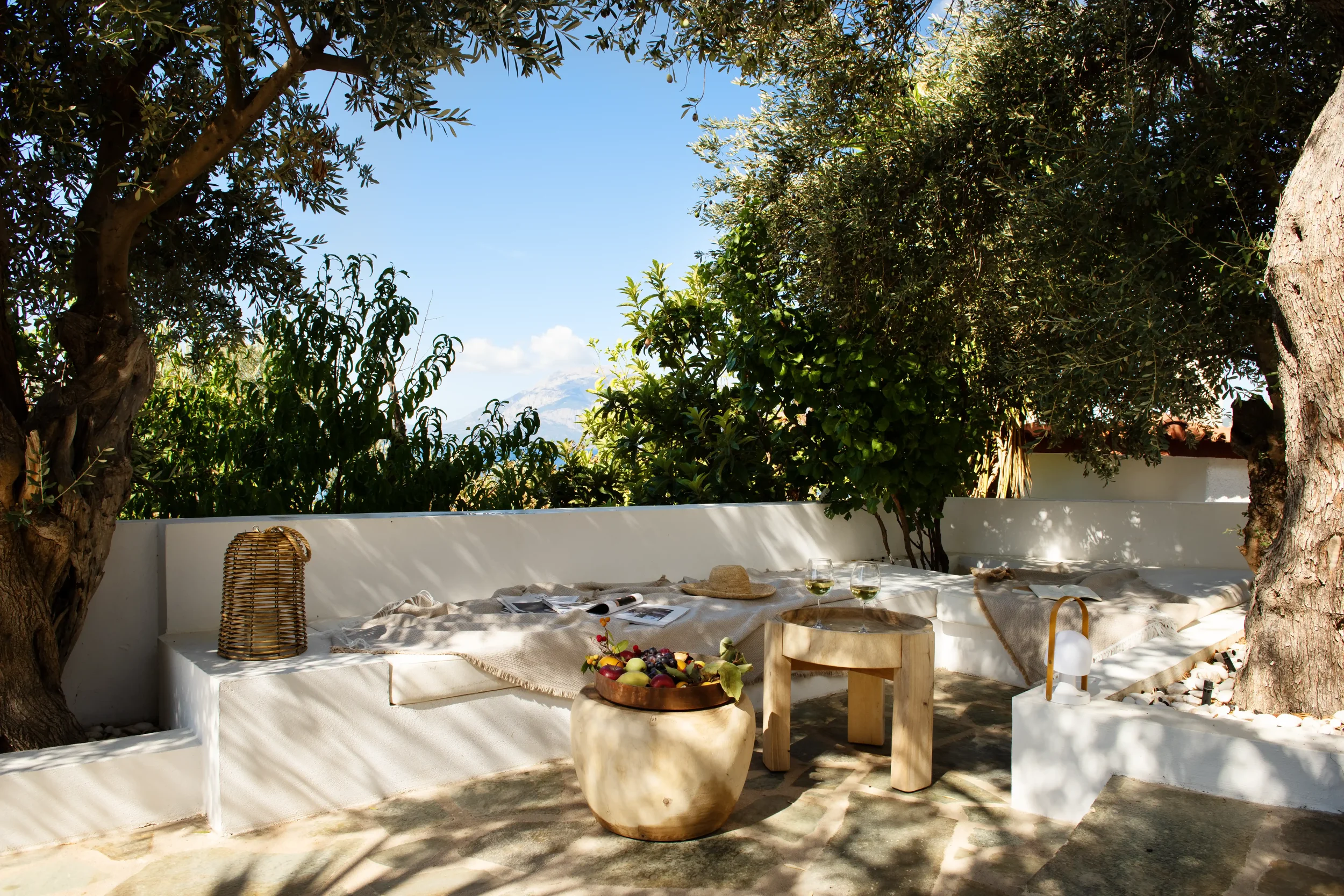 Outdoor seating area with white built-in bench, blanket, and table with wine glasses, hat, and magazines, surrounded by trees and greenery under a partly cloudy sky.