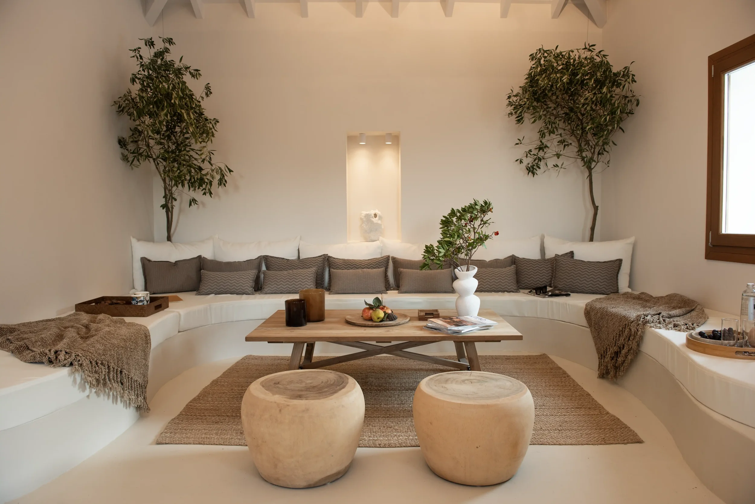 A cozy living room with a curved white sofa decorated with gray pillows, a wooden coffee table with candles, a white vase with green foliage, and two tree-like plants in the background. There's a window on the right side and woven carpets on the floo