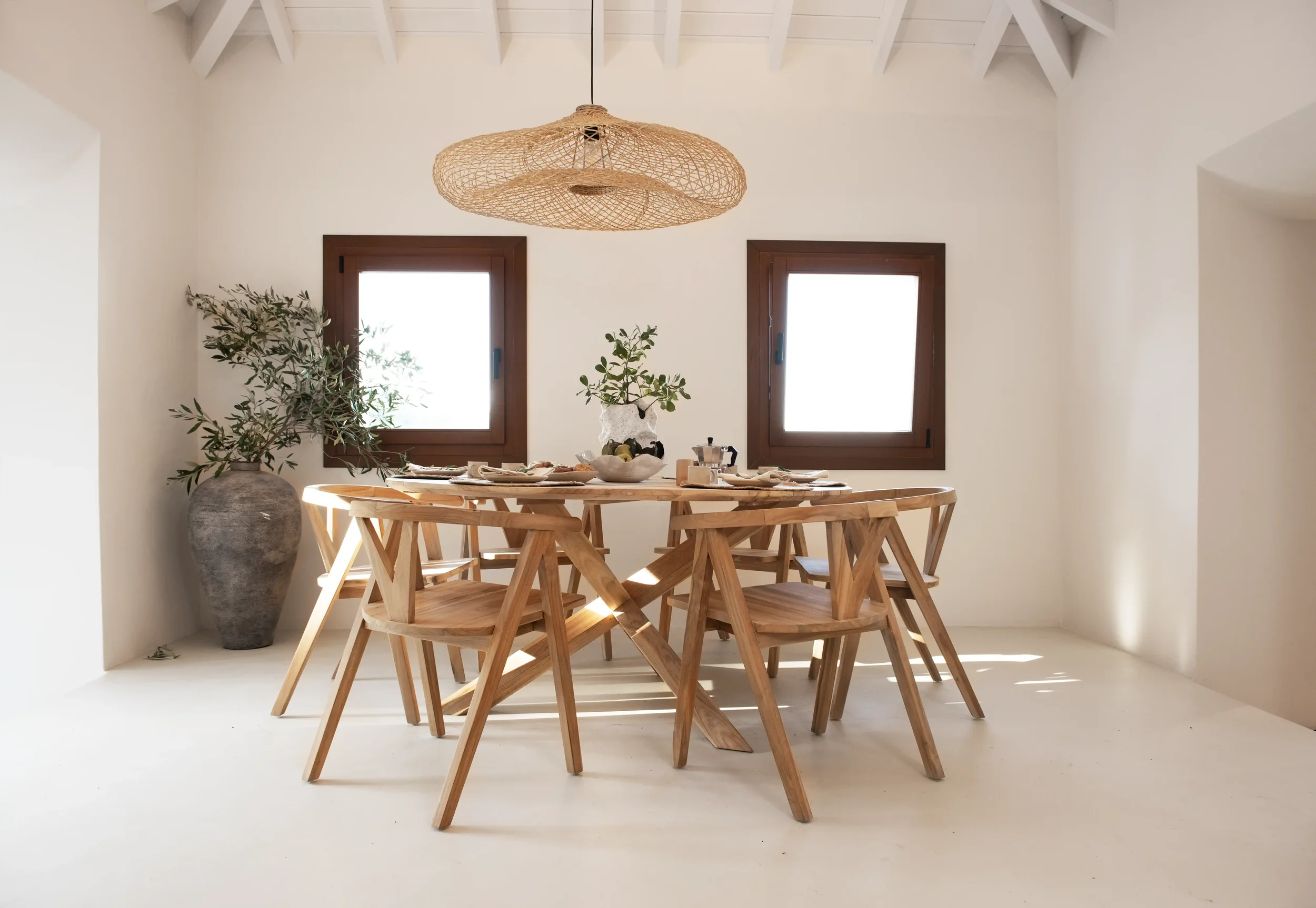 Dining room with a wooden table set for a meal, six wooden chairs, large vase with green plant, two small windows, woven pendant light, white walls, and a white ceiling with exposed beams.