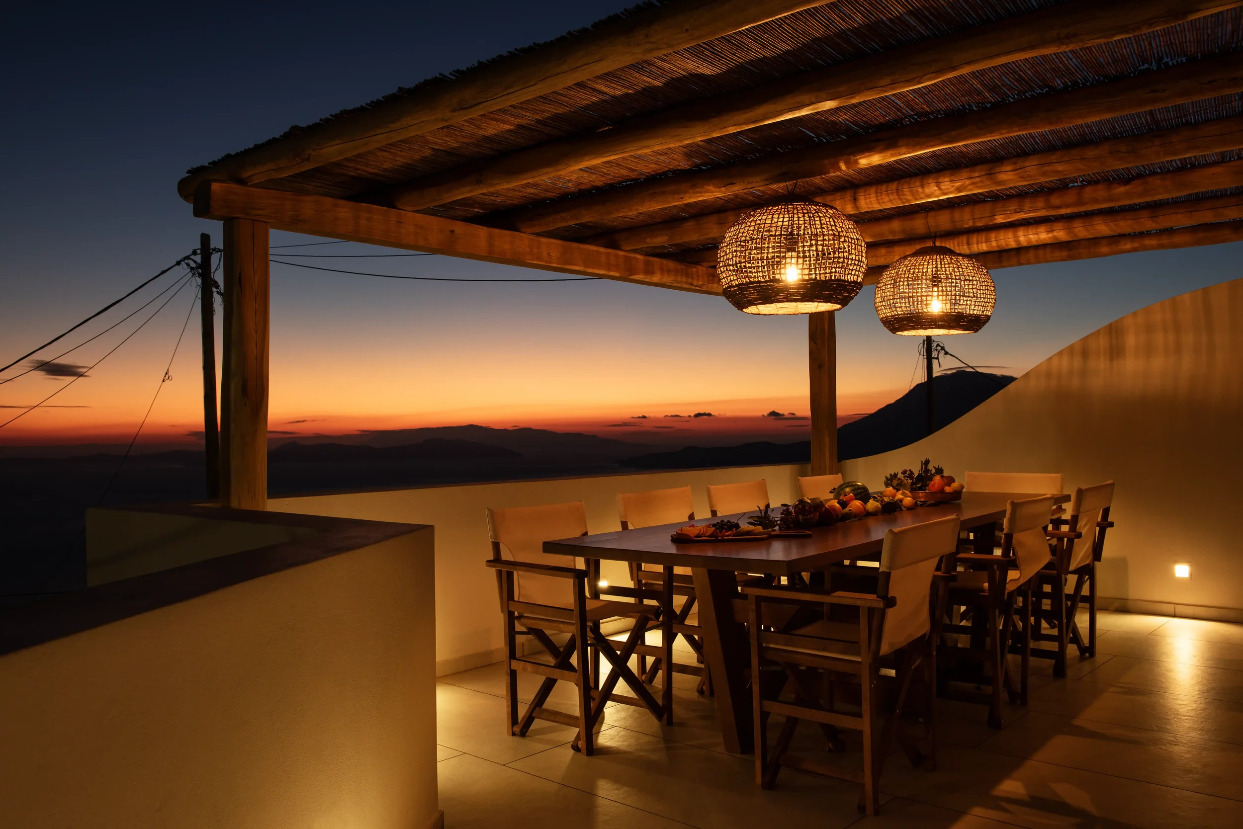 An outdoor dining area with a large wooden table and chairs, decorated with a centerpiece of fruits and vegetables, under a wooden ceiling with hanging wicker lanterns, overlooking a sunset sky with mountains in the distance.