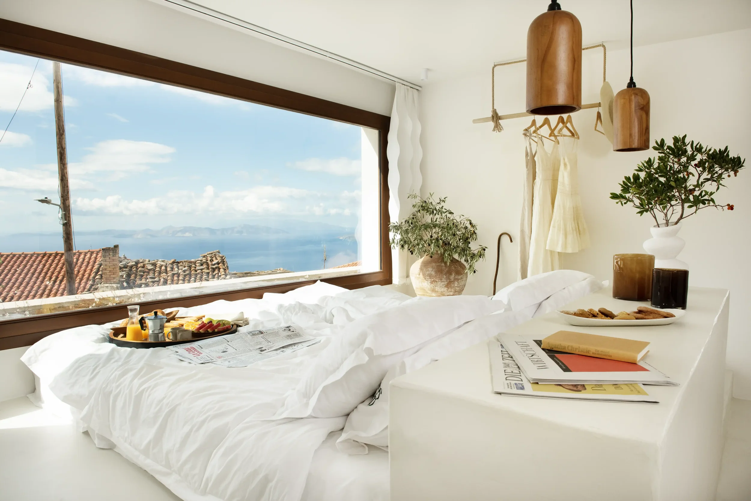 A cozy bedroom with a large window overlooking rooftops, the ocean, and mountains. The bed has white linens with a breakfast tray, newspapers, and a magazine. There are potted plants and hanging wooden lights in the room.