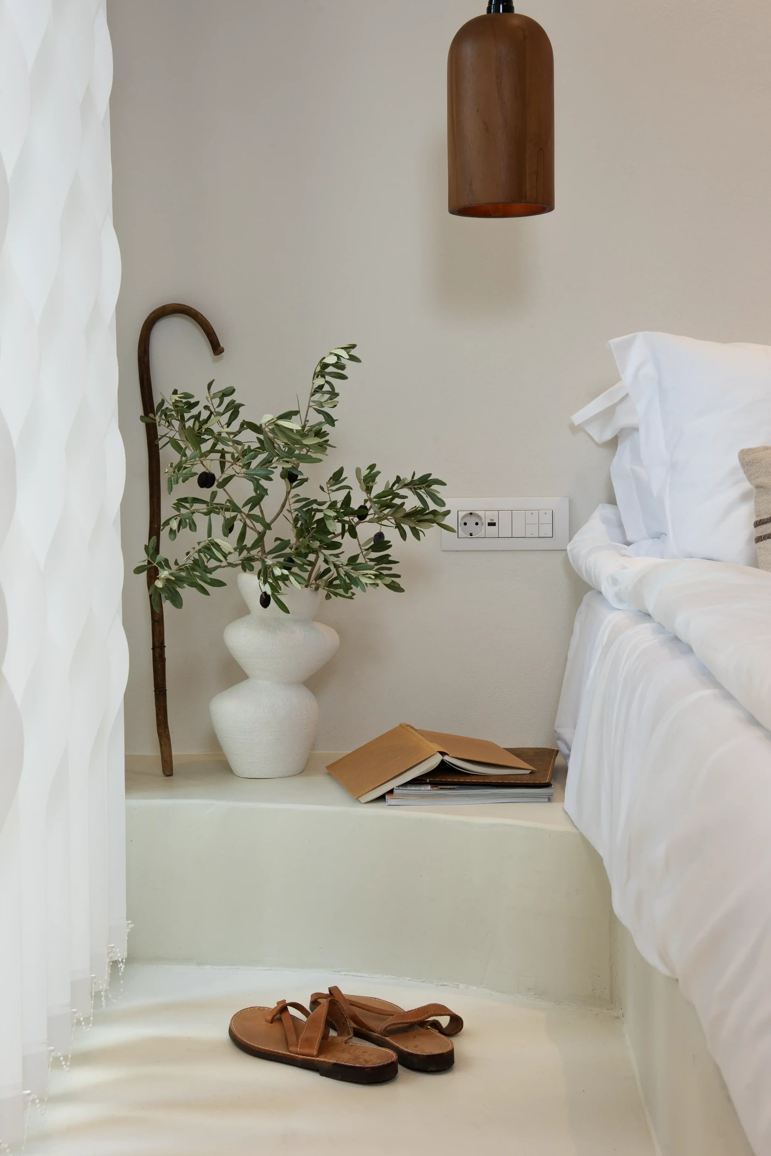 Interior of a bedroom corner with a white bed, a white ceramic vase with greenery, a wooden umbrella stand, a stack of books, a pair of sandals, and a wall socket.