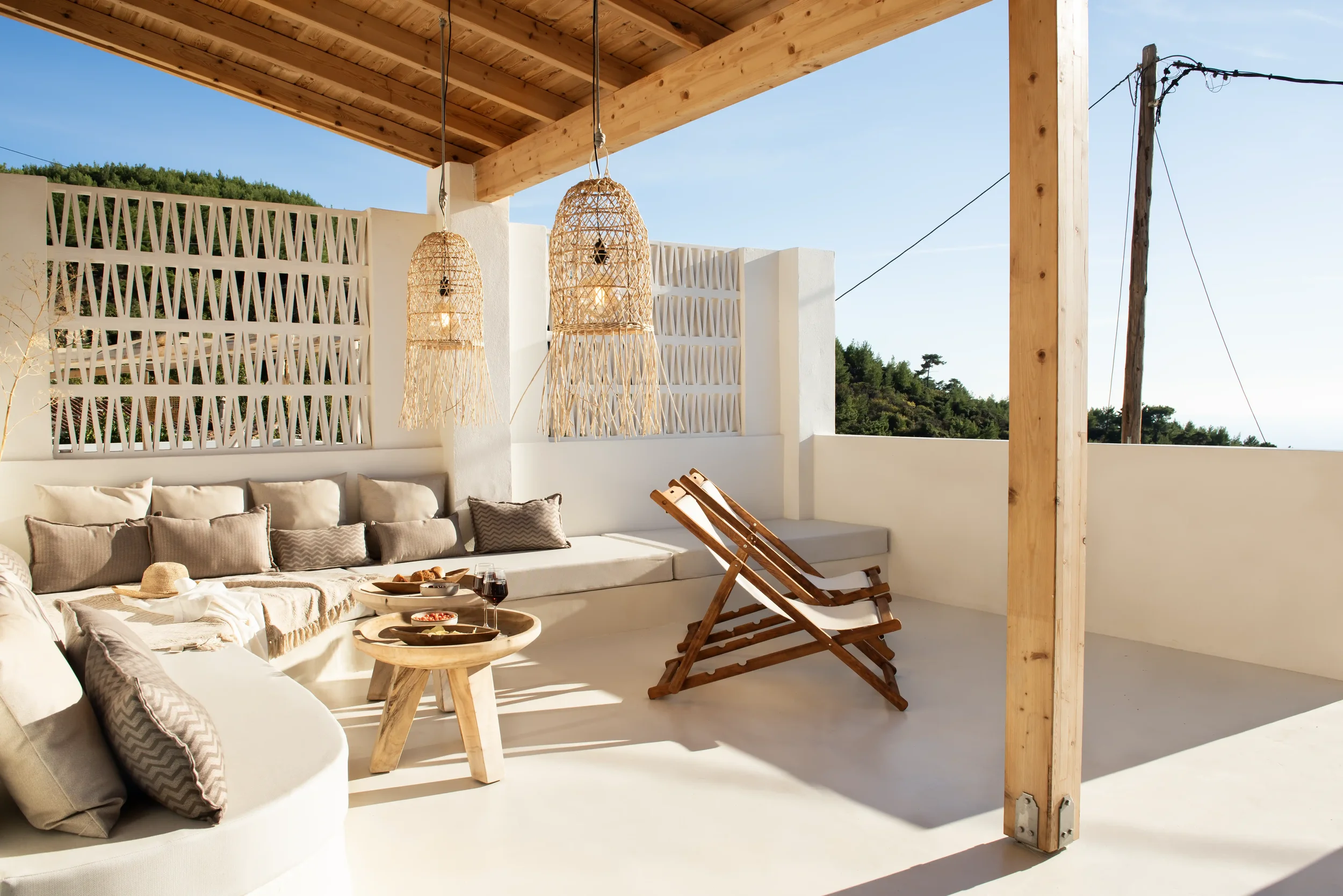 Sunlit outdoor terrace with white walls, beige cushioned seating, wooden lounge chairs, small wooden table set with food and drinks, and hanging wicker pendant lights under a wooden roof, overlooking green hills.