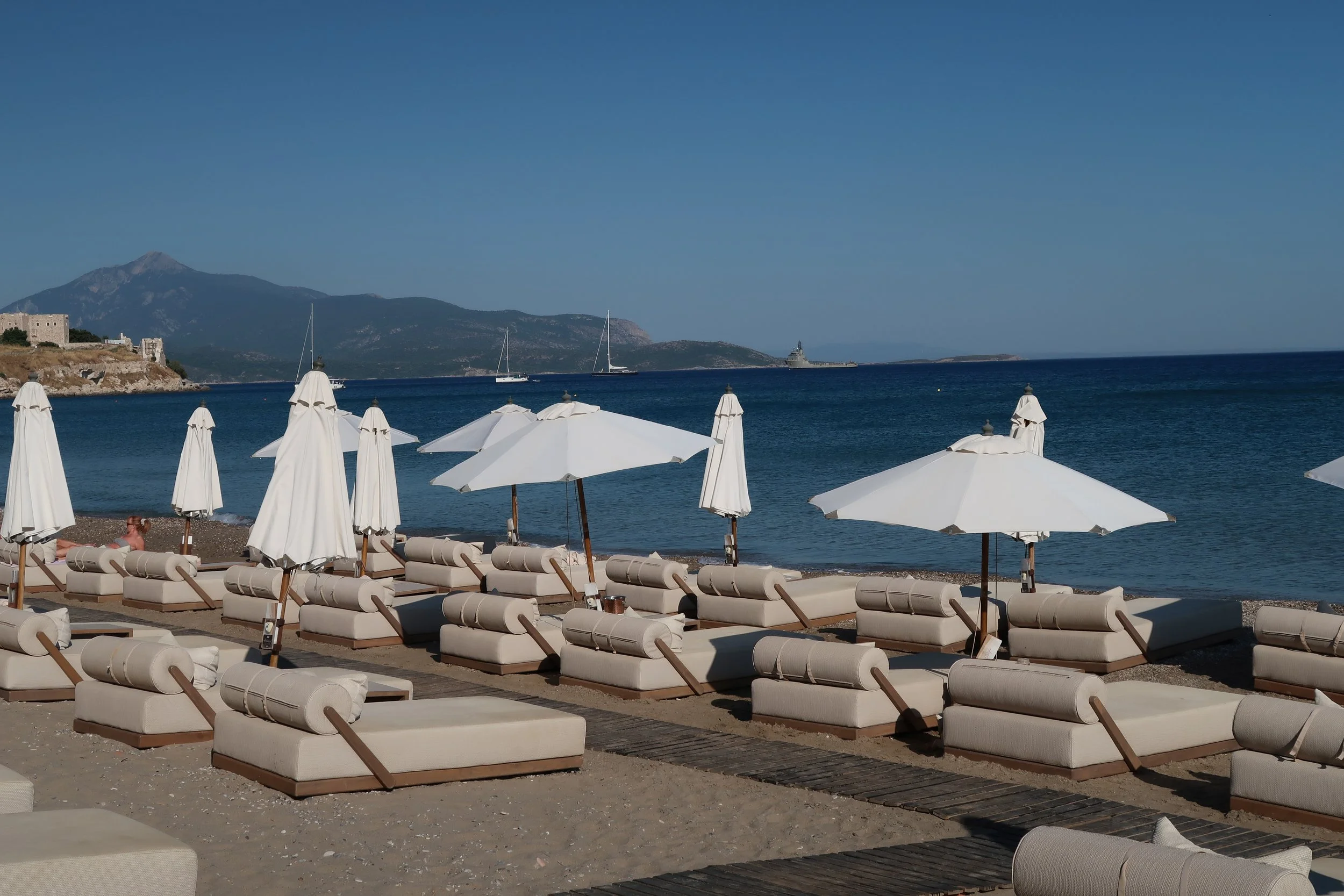 Empty beach with white lounge chairs and umbrellas facing the water, with mountains and sailboats in the background.