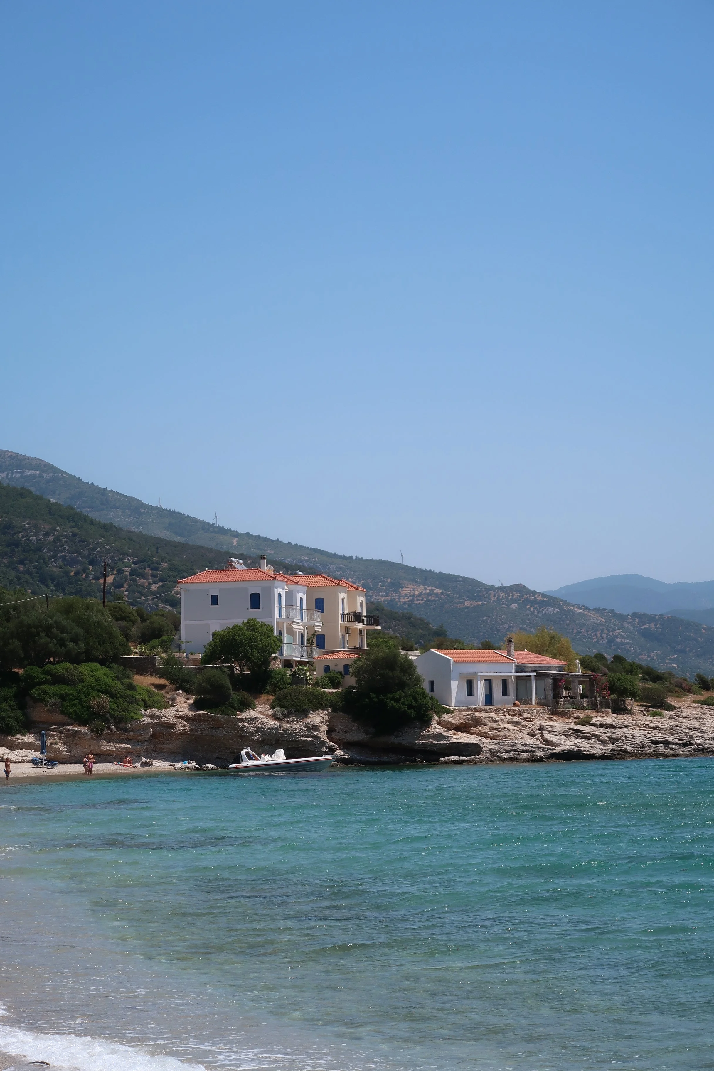 Beach with houses on rocky coast, boat near shore, hills in background, clear blue sky.