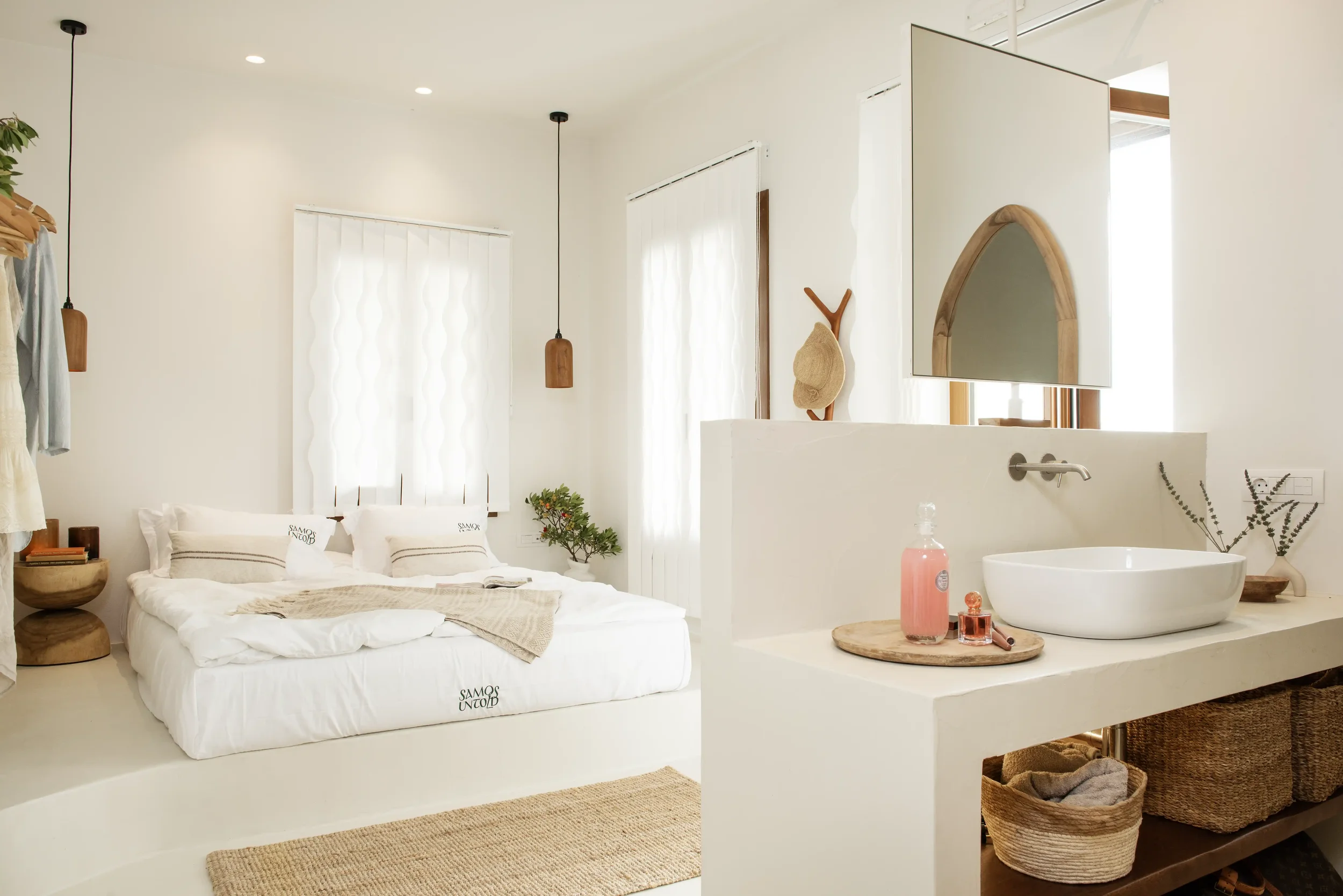 A bright, minimalist bedroom with a white bed, wooden accents, hanging pendant lights, a small plant, and a white bathroom vanity with a sink, mirror, and decor items.