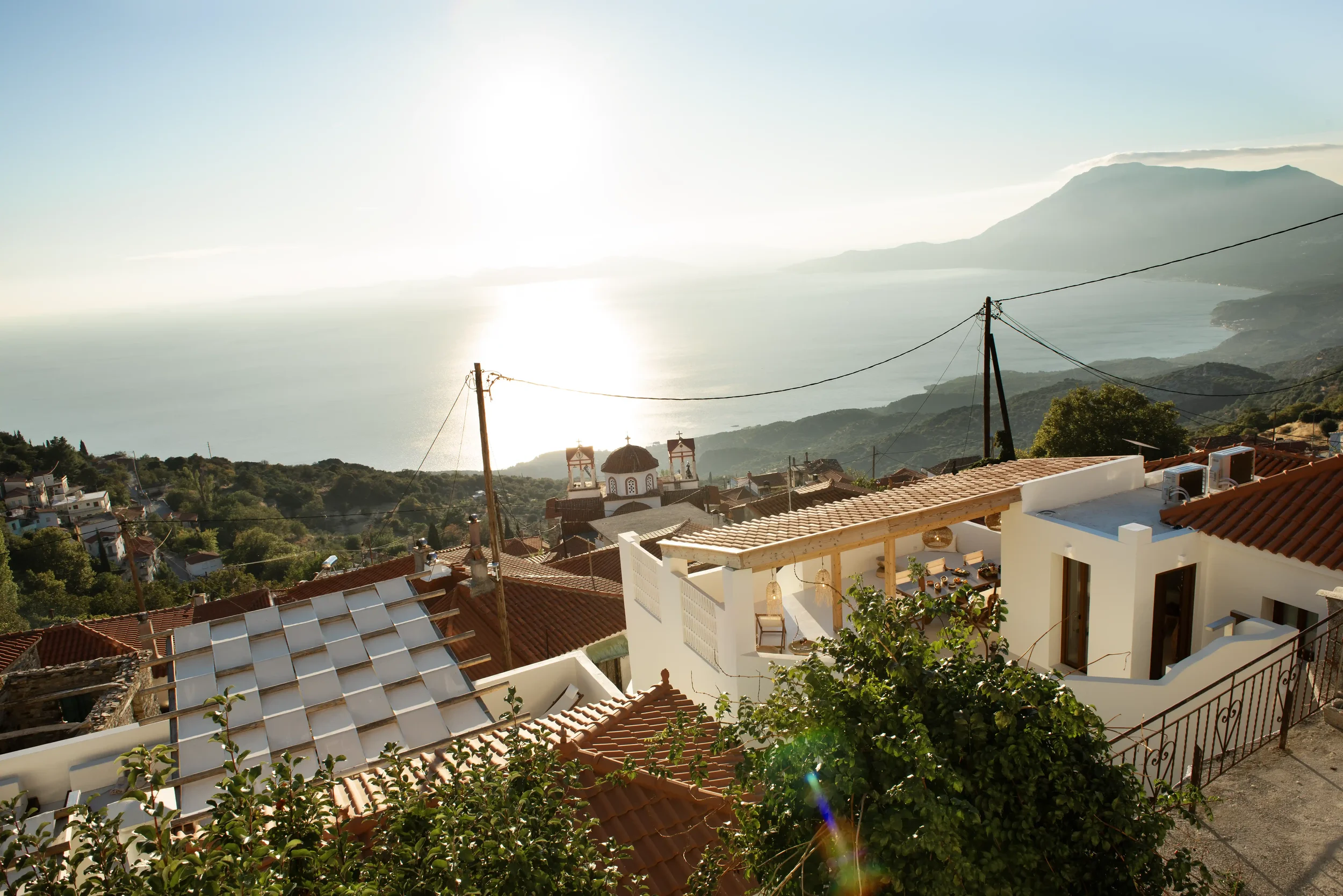 View of a coastal town with red-roofed buildings, a church with domes, and mountains in the background during sunset over the ocean.