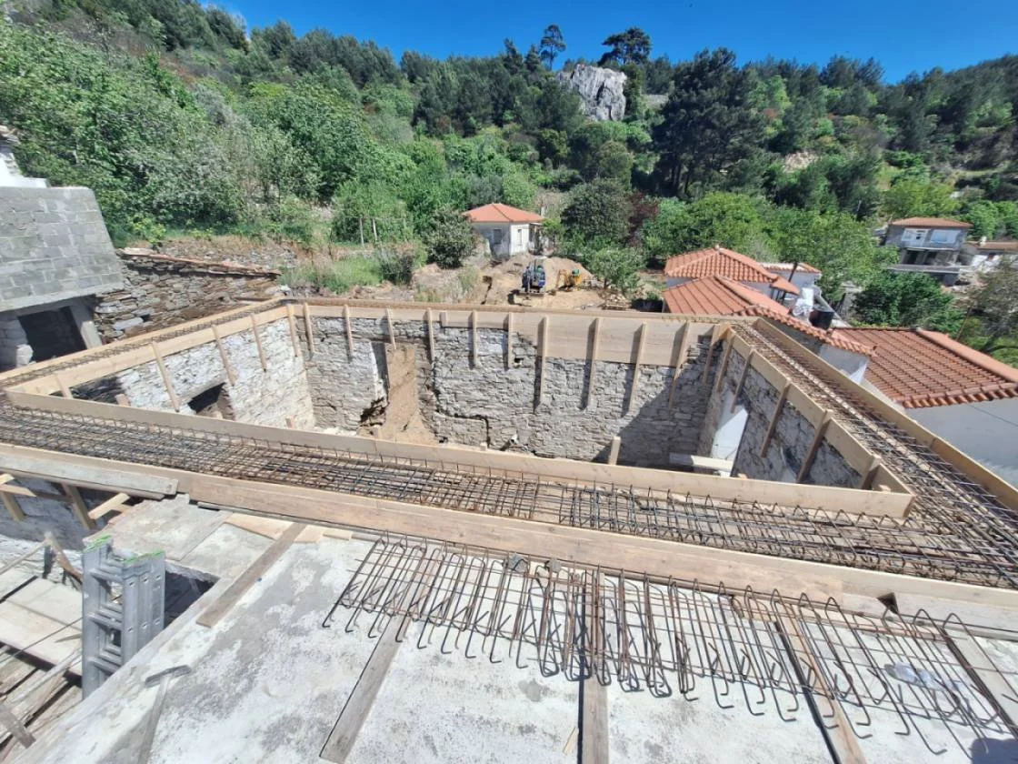 Construction site with a partially built stone structure, rebar reinforcement, and wooden formwork, surrounded by green trees and neighboring houses with red-tiled roofs.