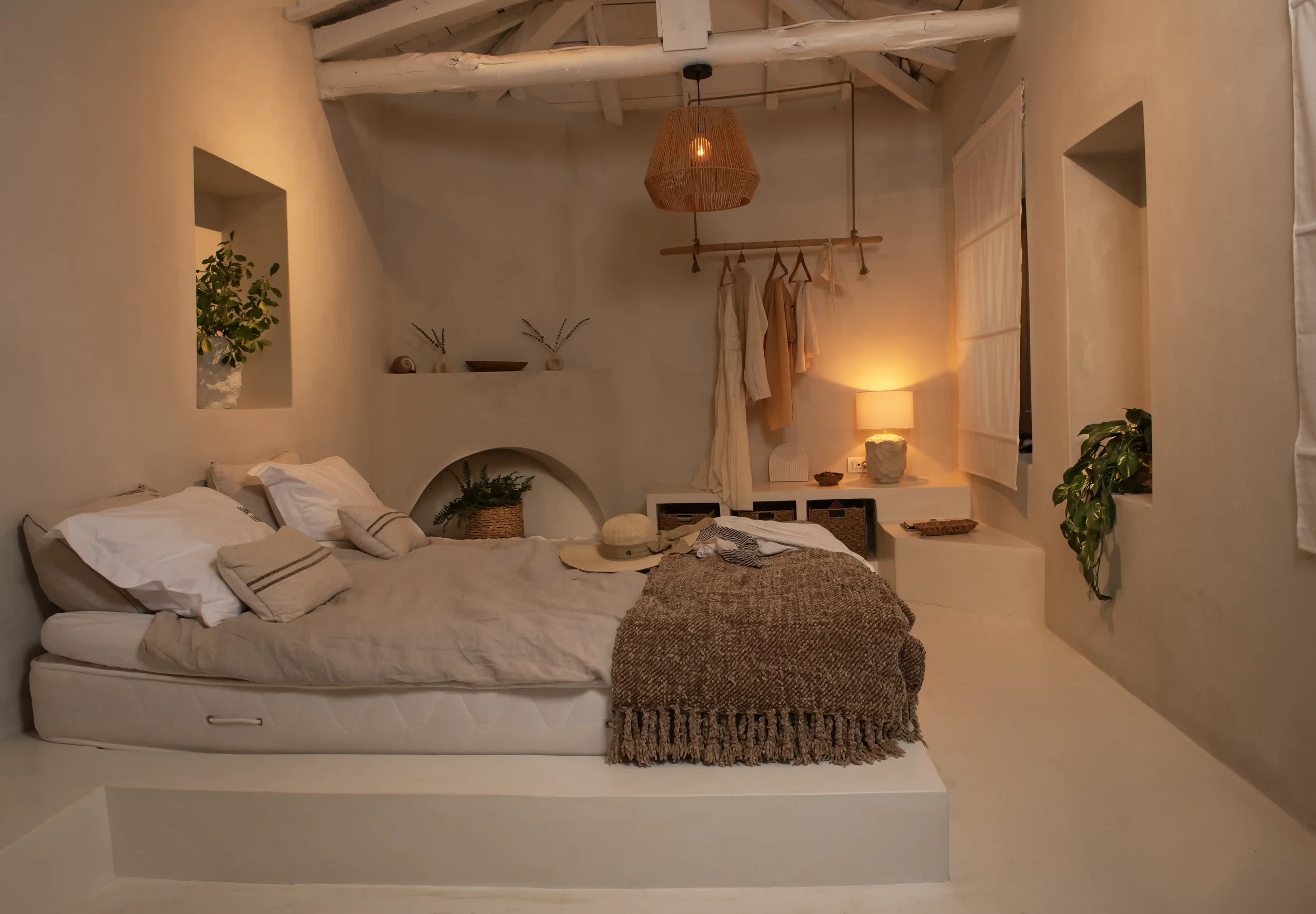 A cozy bedroom with a white bed, beige bedding, and pillows. There is a straw hat on the bed and a textured blanket. The room has soft, warm lighting from a bedside lamp. Plants are placed in wall niches and on a shelf. Neutral tones and natural mate