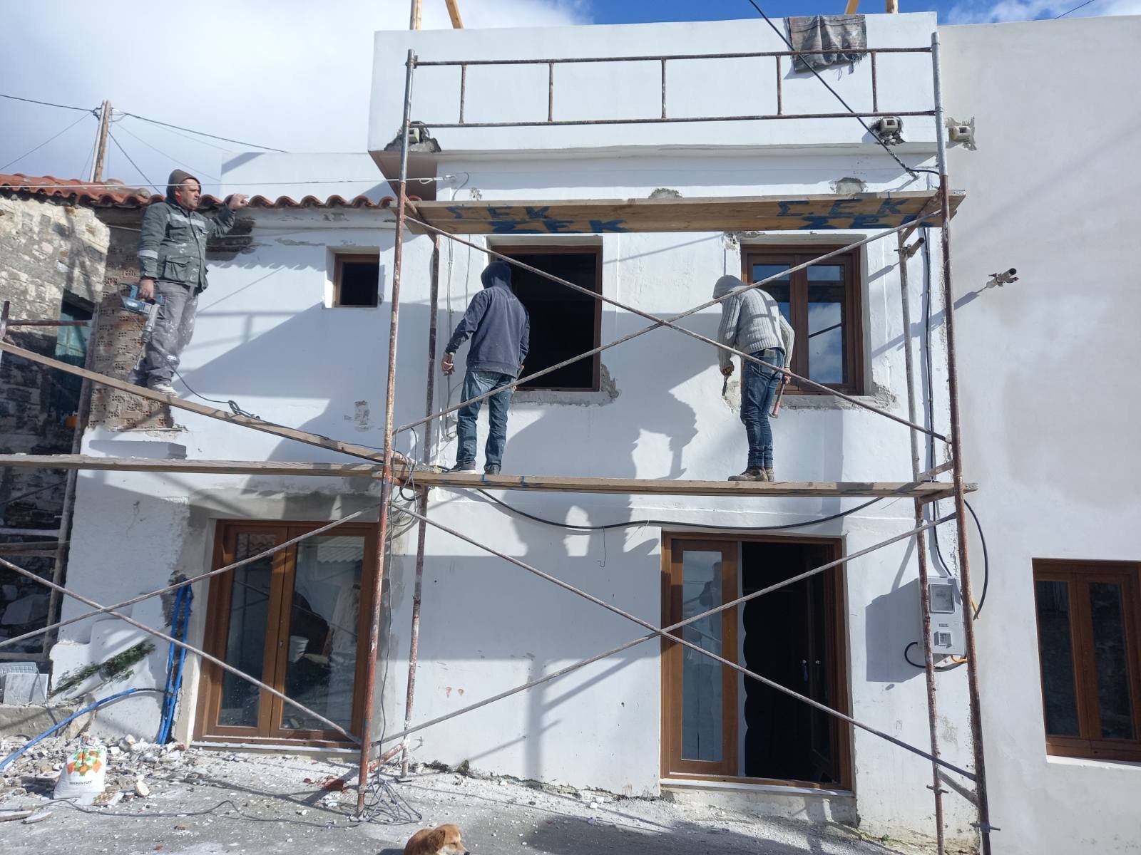 Three workers are building or renovating a white residential building, standing on scaffolding in front of the house with a door and windows. One worker is on the left holding a trowel, and two others are on the right, working near windows. The build