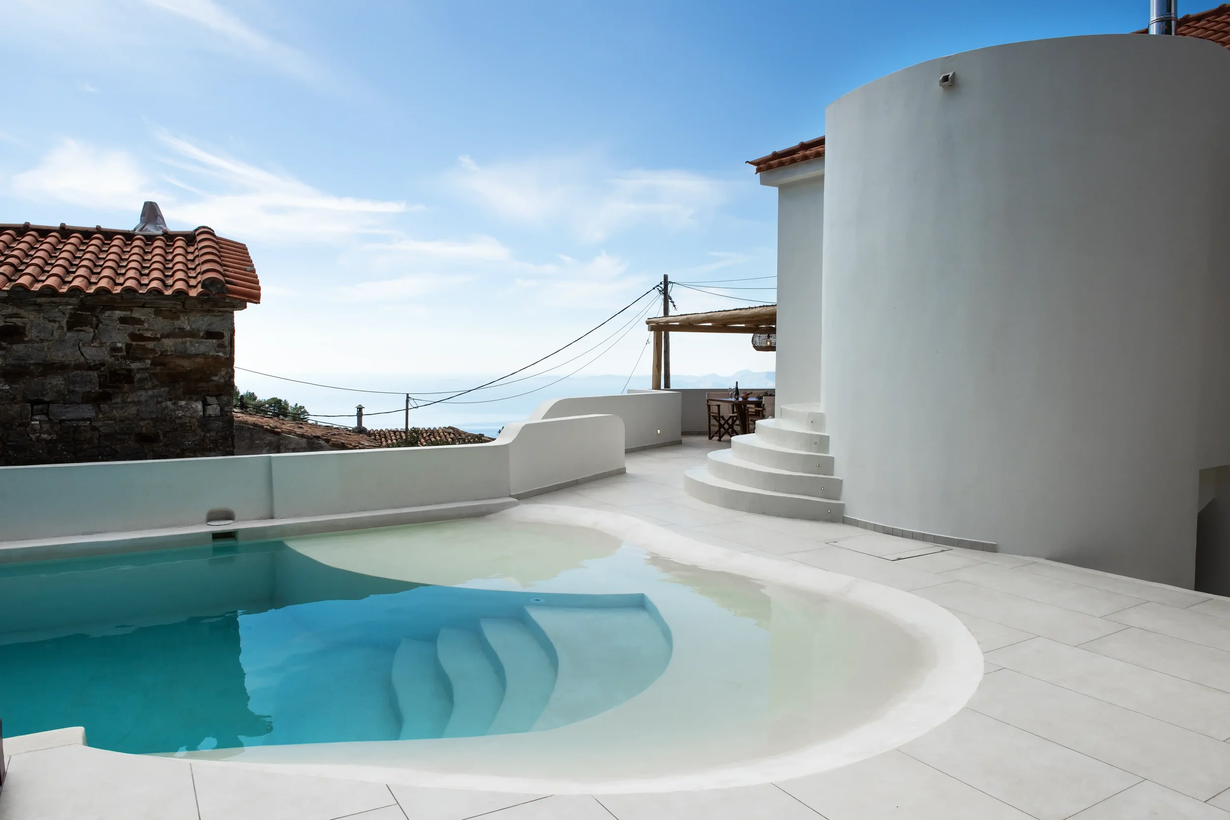 Modern white terrace with a small pool, outdoor table and chairs, and distant ocean view under blue sky with clouds.
