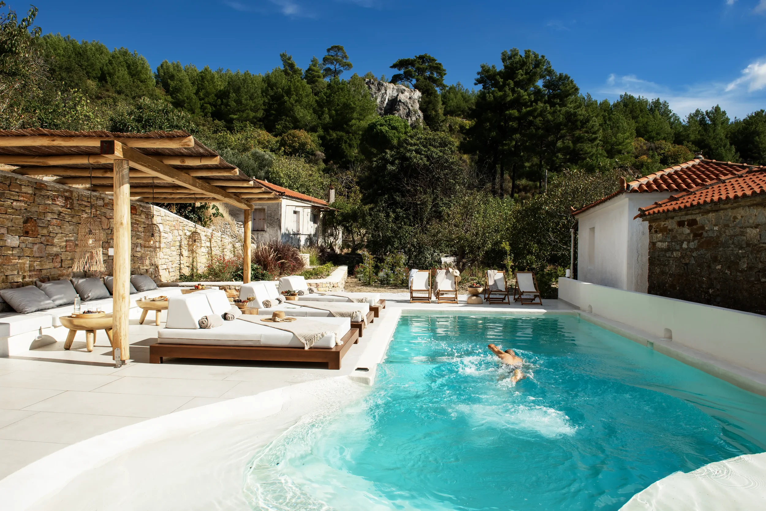 A person swimming in a turquoise outdoor pool at a sunny villa with lounge chairs, a stone wall, a wooden pergola, and lush green trees in the background.