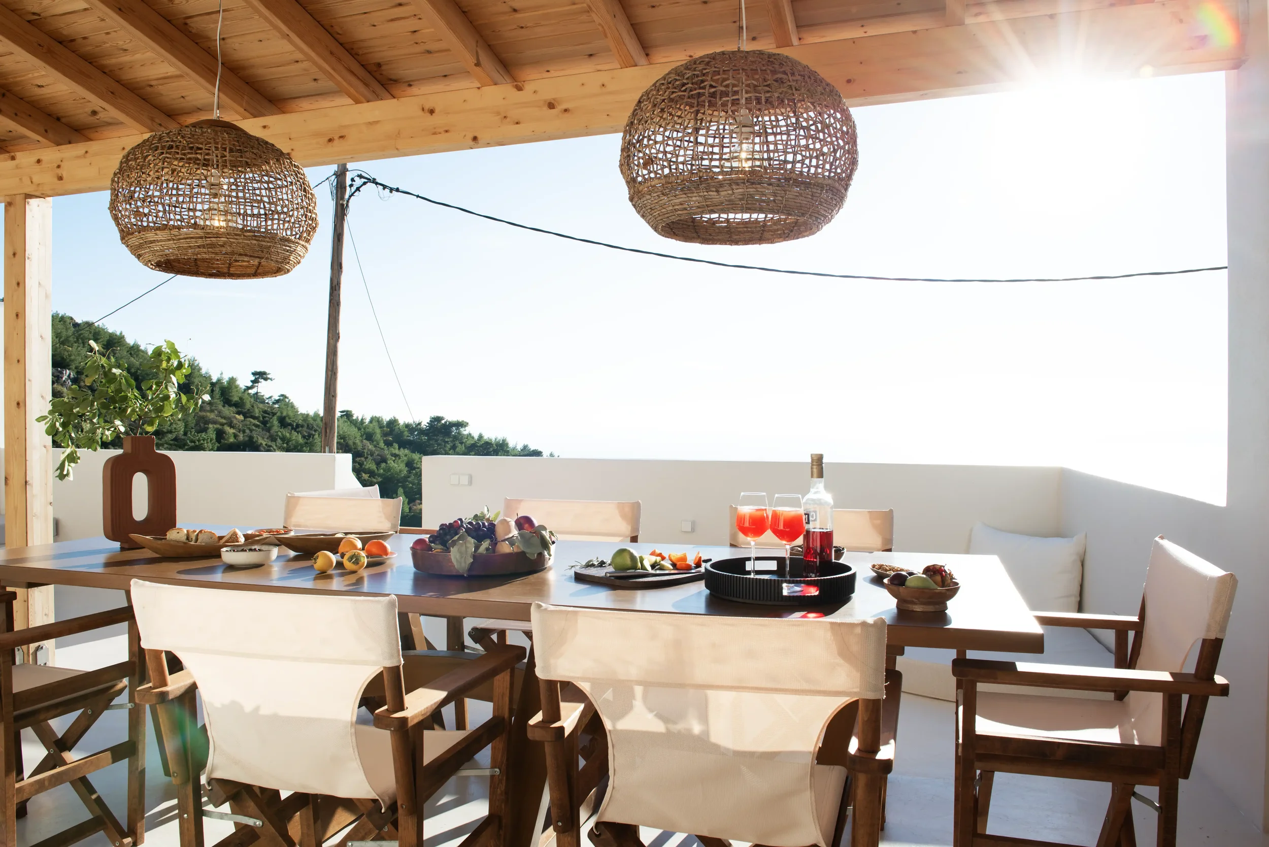 Outdoor terrace with wooden ceiling, hanging wicker light fixtures, a dining table set with glasses of red drink, bowls of fruit, and food, surrounded by wooden chairs and a white cushioned bench, with greenery and a bright sky in the background.